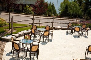 Un patio al aire libre con mesas redondas y sillas de mimbre, rodeado por un borde de piedra y follaje.