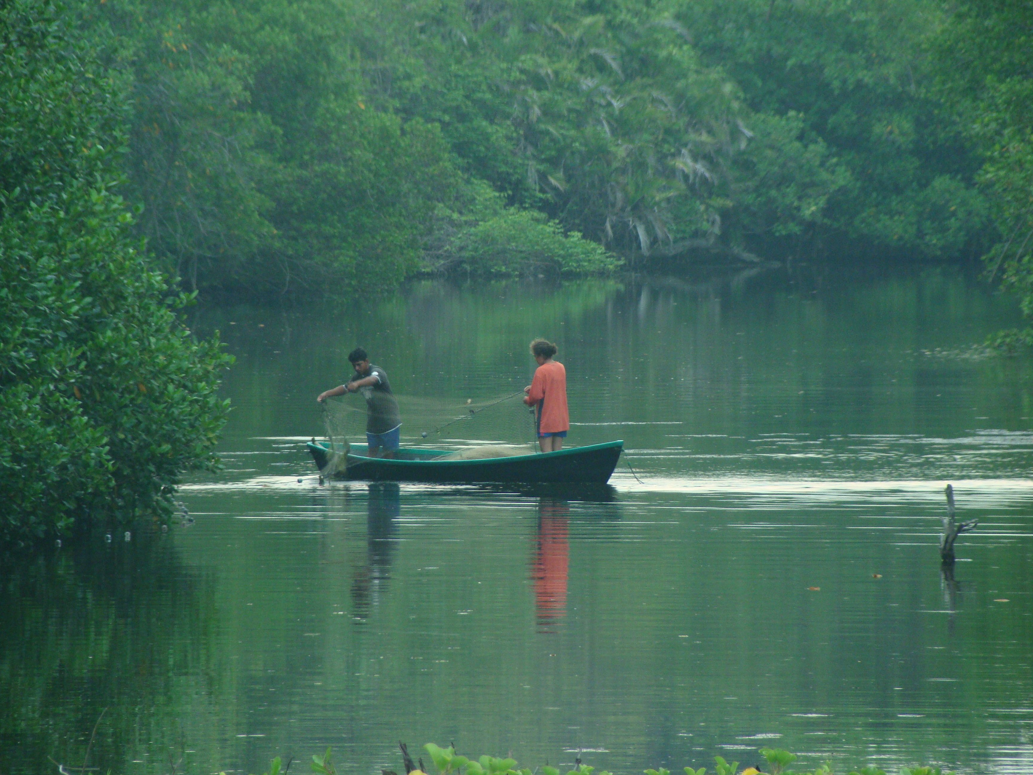 A calm river with two people on a small boat, surrounded by dense greenery.