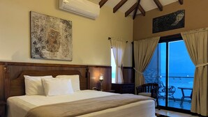 Une chambre d’hôtel avec un grand lit, une table de chevet, une lampe, un tableau et une vue sur la mer, visible à travers une porte vitrée.