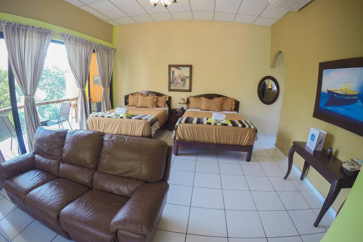 Ein Hotelzimmer mit zwei Betten, einem Sofa, einem Balkon mit Vorhängen, einem Gemälde und einem gerahmten Bild an der Wand.