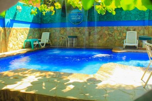 Ein Schwimmbecken mit klarem, blauem Wasser, umgeben von Steinmauern und blauen und grünen Dekorationselementen. In der Nähe des Pools befinden sich Liegestühle und ein kleiner Tisch.