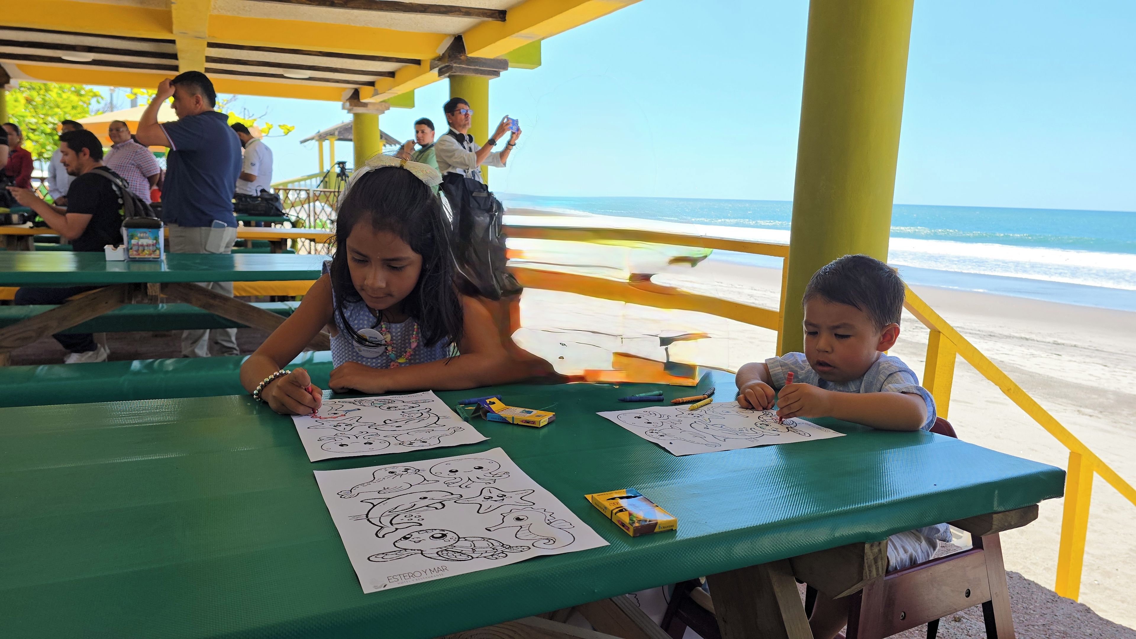 Children drawing at a beachside table with a view of the ocean.
