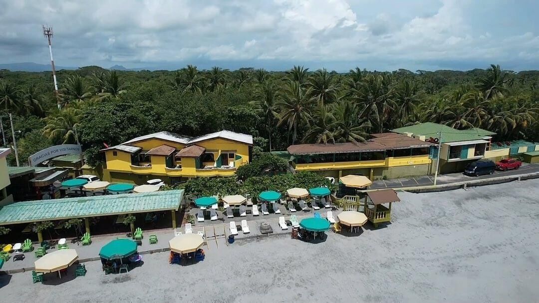 A beachside resort with multiple yellow buildings, outdoor seating areas, and a view of palm trees.