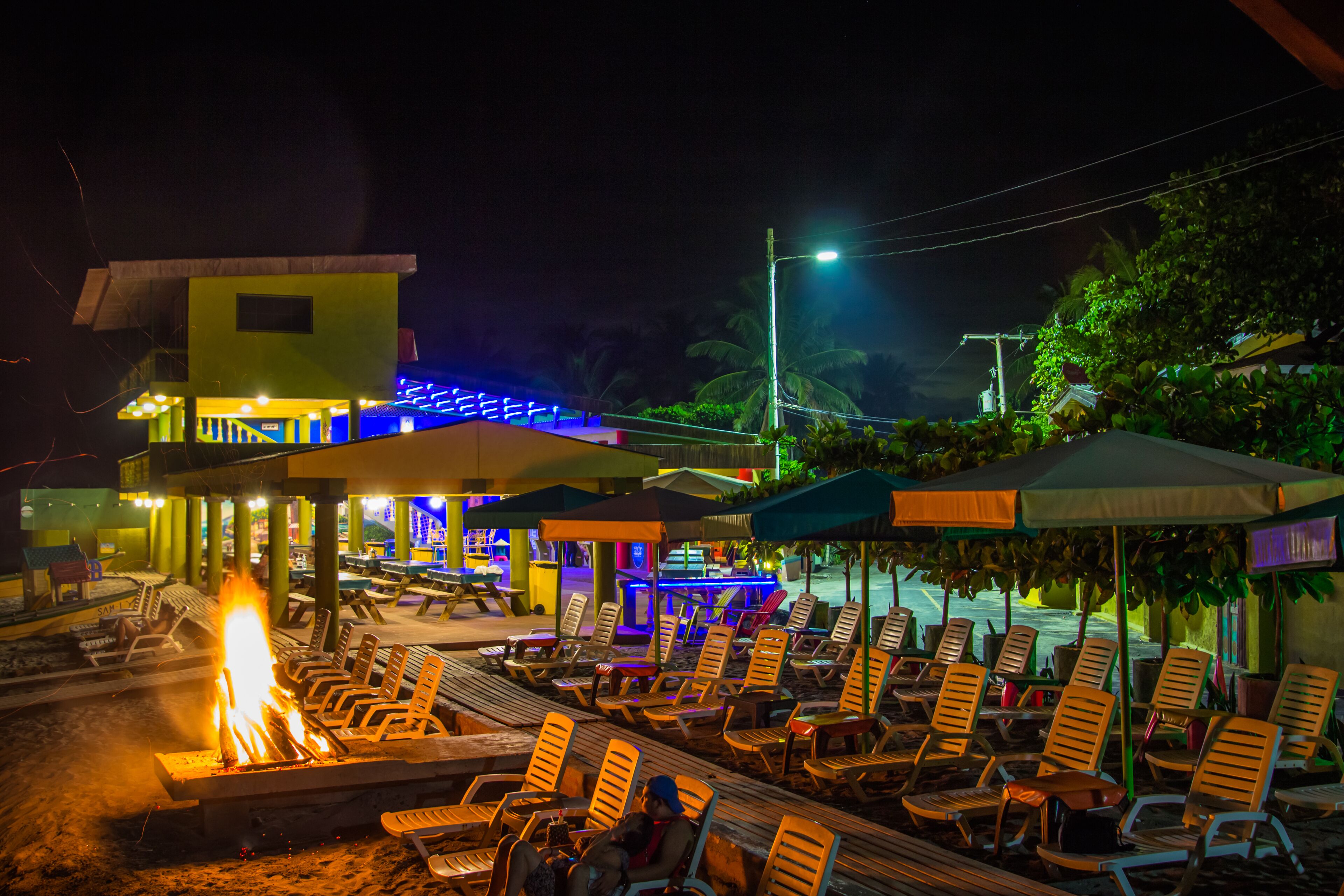 A beachside lounge with lounge chairs and a fire pit, surrounded by umbrellas and a building with illuminated blue lights.