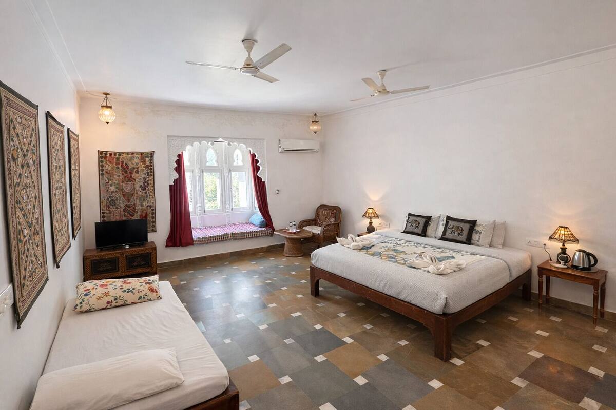 Une chambre avec un grand lit, un plus petit lit, une télévision, une fenêtre avec des rideaux, un ventilateur de plafond et un élément décoratif mural.