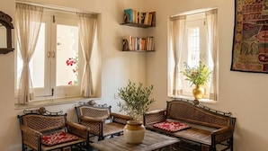 A room with four wicker chairs, a wooden coffee table, and two windows with curtains. There are bookshelves with books and a large tapestry on the wall. - Hibiscus Heritage - A Lake View Hotel in Udaipur (Udaipur)