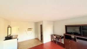 A room with a wooden floor, a desk with a chair, a television on a wooden stand, and a white wall with a mirror and an air conditioning unit. - Portland Value Inn & Suites Near PDX Airport (Portland)