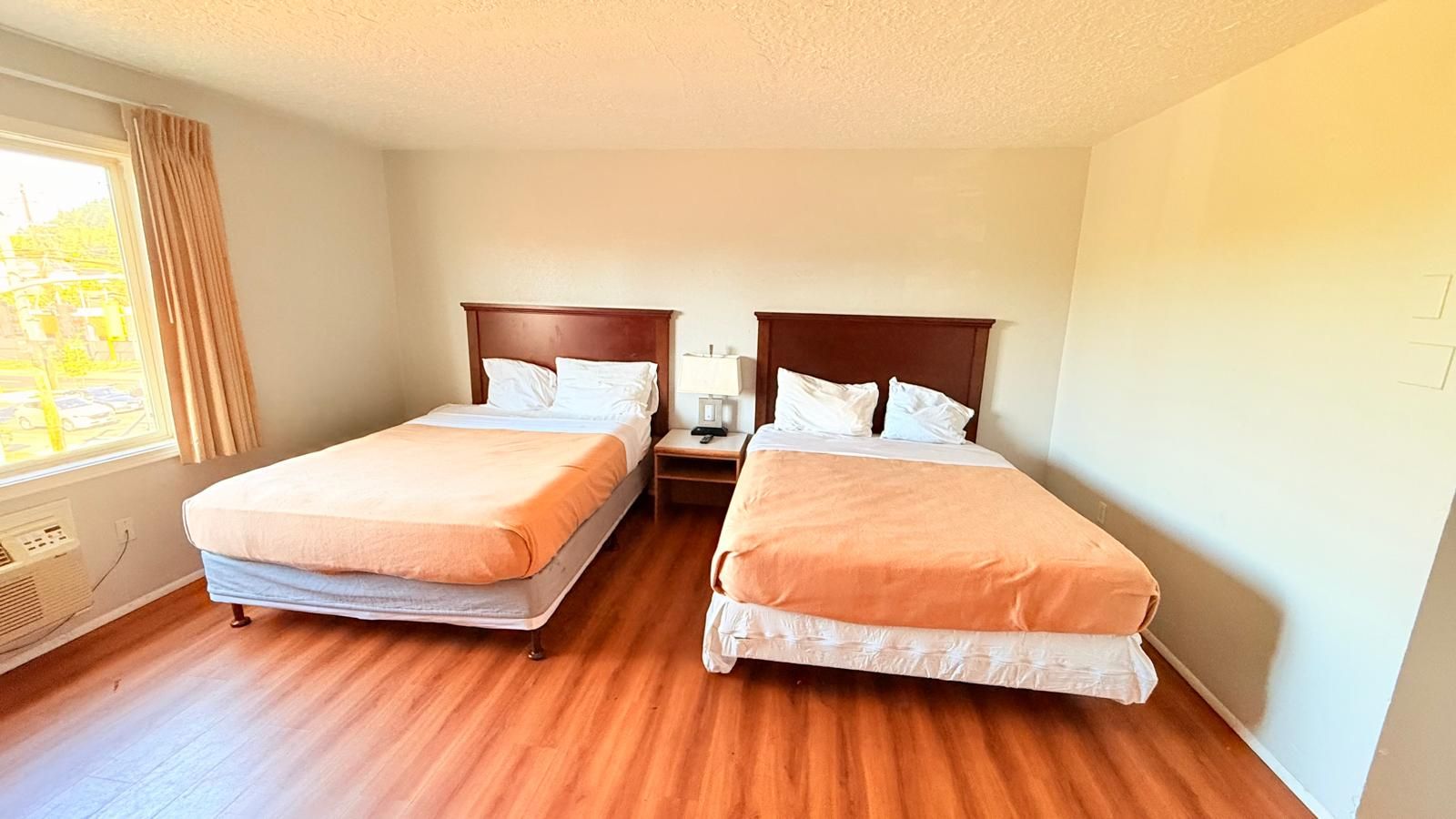 Habitación de hotel con dos camas individuales, piso de madera, ventana con cortinas y una mesita de noche con lámpara.
