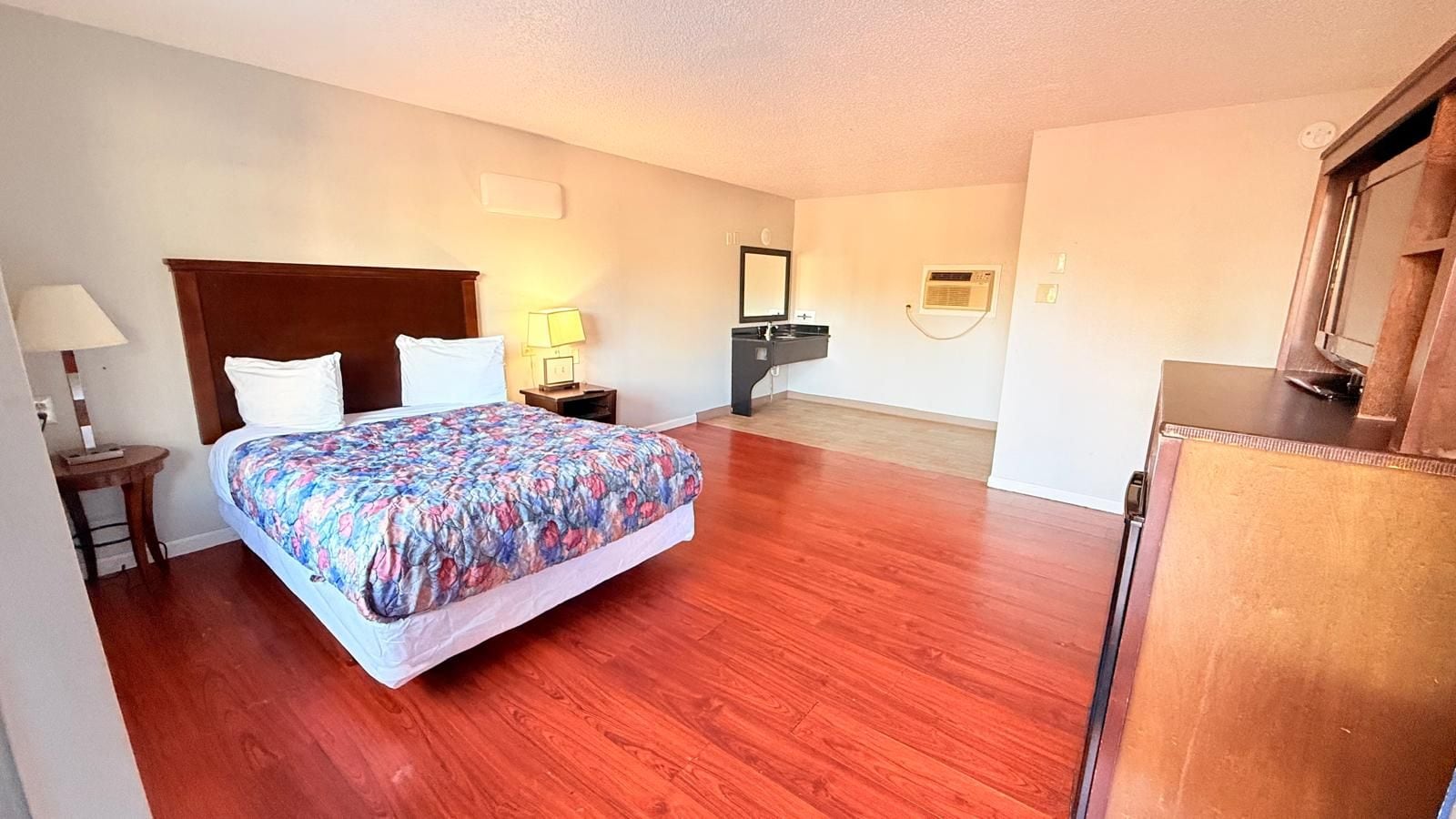 Habitación de hotel con cama, mesita de noche, lámpara y baño con espejo y lavamanos.