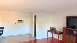 A room with a wooden floor, a flat-screen TV on a wooden stand, a small table with a microwave, and a door leading to another room.