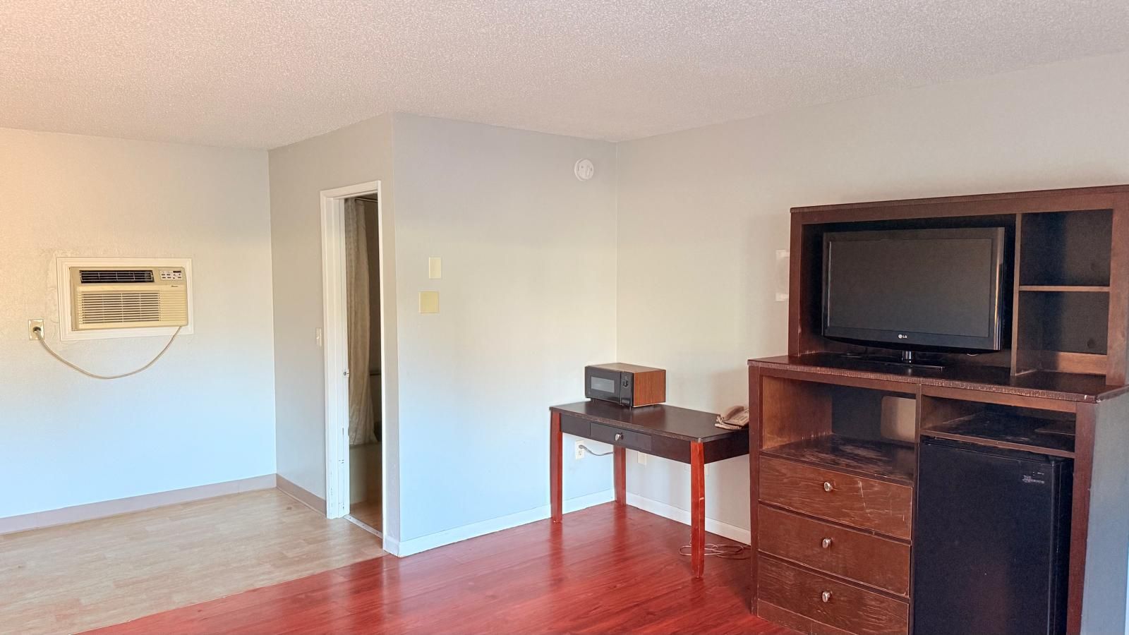 A room with hardwood flooring, a wooden desk with a computer, a television on a wooden stand, and a door leading to another room.