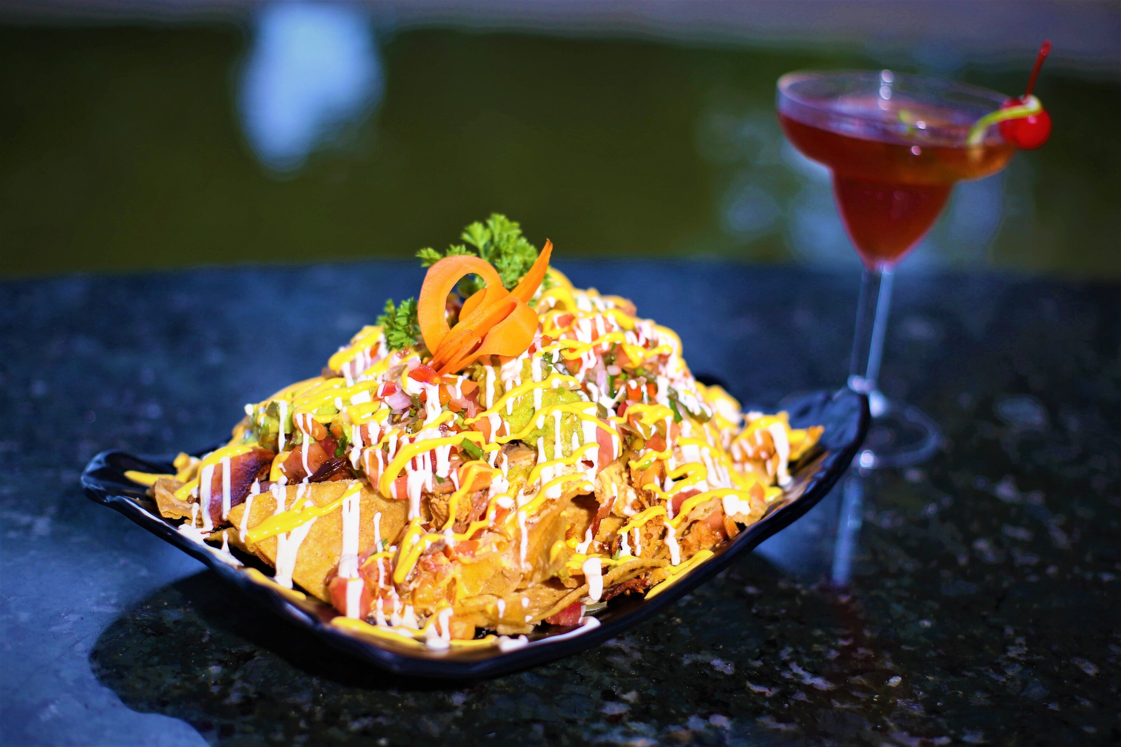 Une assiette de nachos recouverte de différentes sauces et un cocktail décoré d’une cerise.
