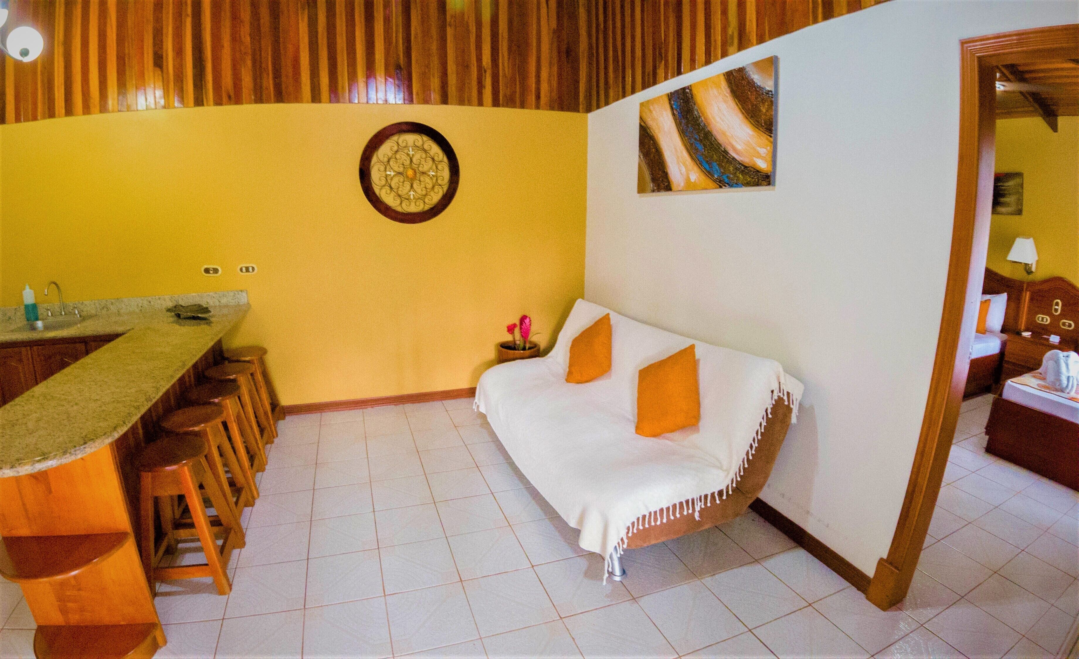 Una habitación compacta con kitchenette, un sofá con cojines naranjas, un reloj decorativo de pared y una pequeña mesa con un florero.