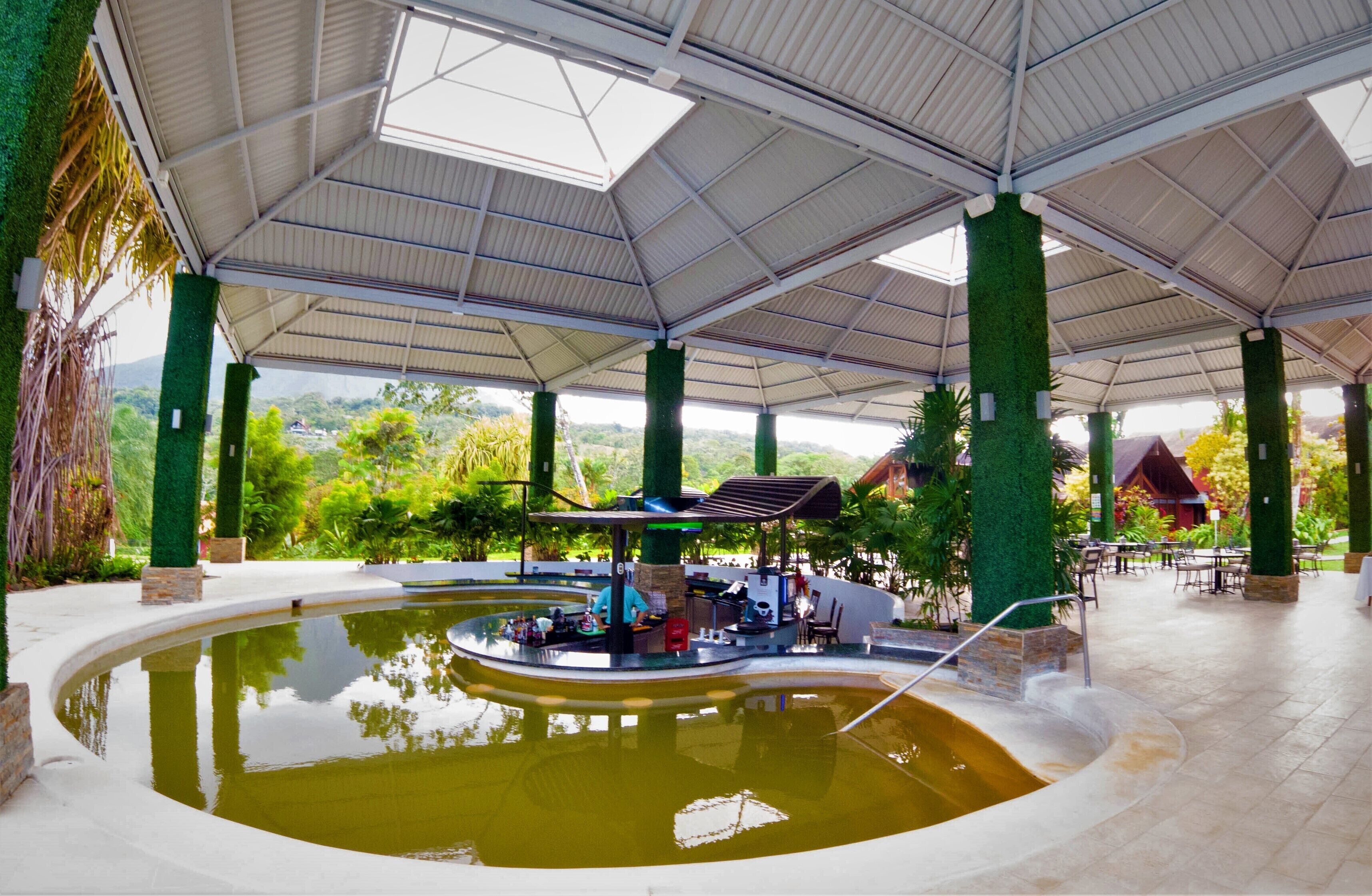An indoor pool area with a central bar and seating, surrounded by greenery and a covered roof structure.