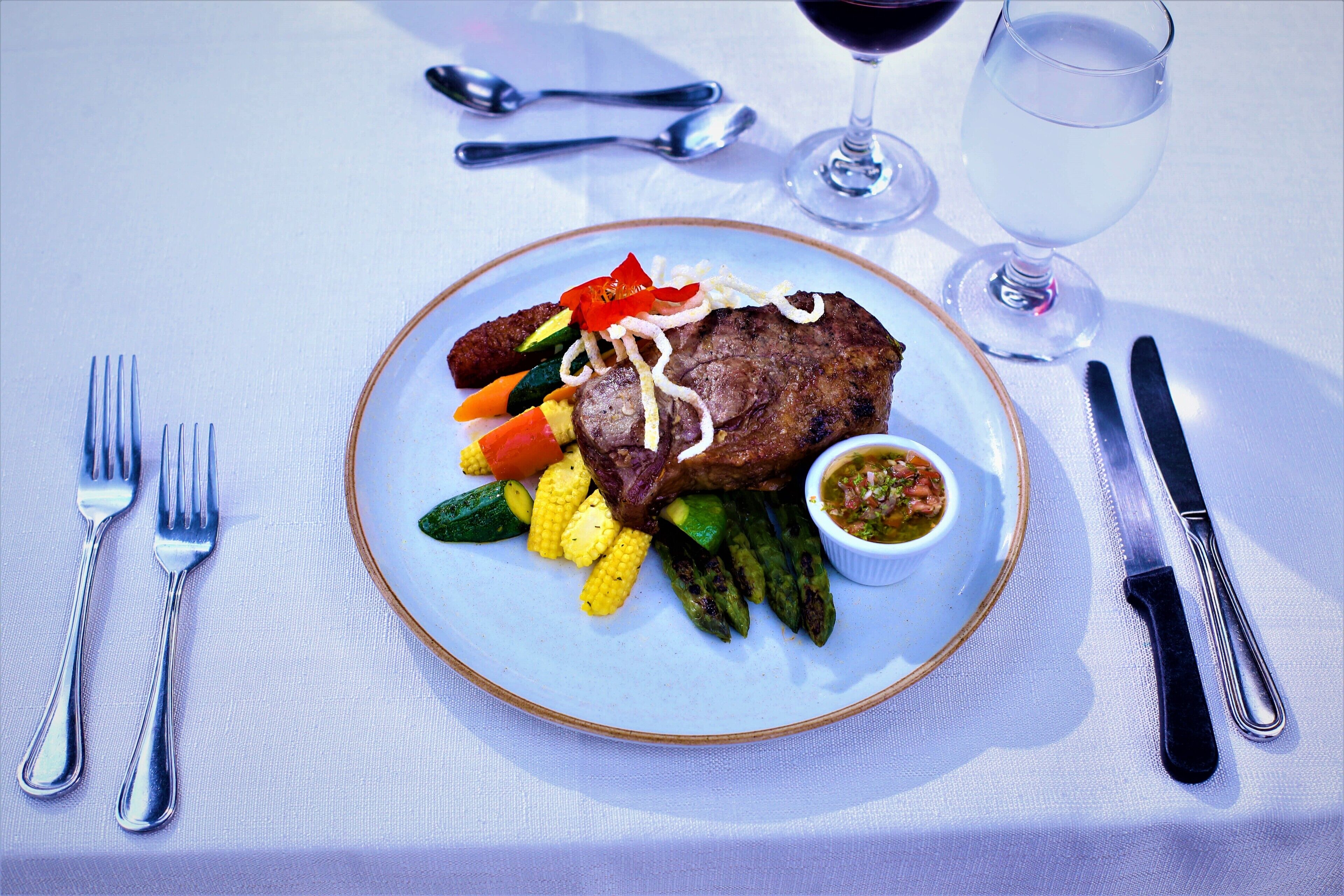 Un piatto di carne con bistecca, verdure e salsa, accompagnato da posate e un bicchiere di vino rosso su un tavolo.