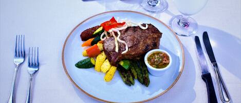 A plate of food with a steak, vegetables, and a sauce, accompanied by cutlery and a glass of red wine on a table.
