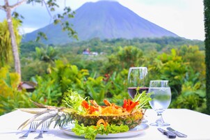 Uma mesa com prato de frutos do mar, copos de vinho e talheres, com um cenário de vegetação exuberante e montanhas ao fundo.