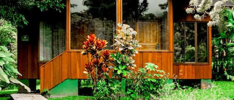 A wooden cabin with large windows, surrounded by lush greenery and flowering plants.