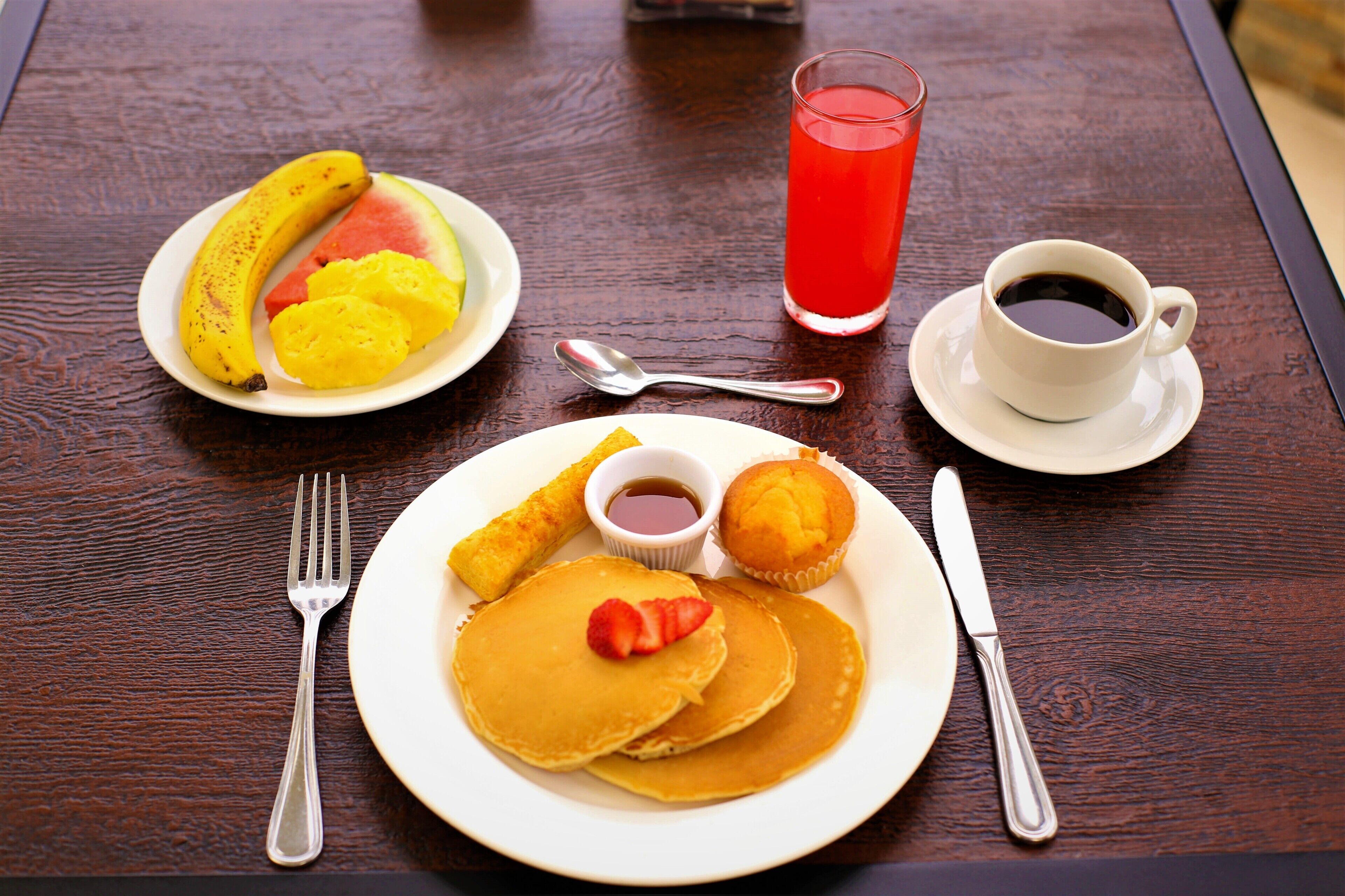 A breakfast setting with pancakes, fruit, a muffin, and a cup of coffee on a wooden table.