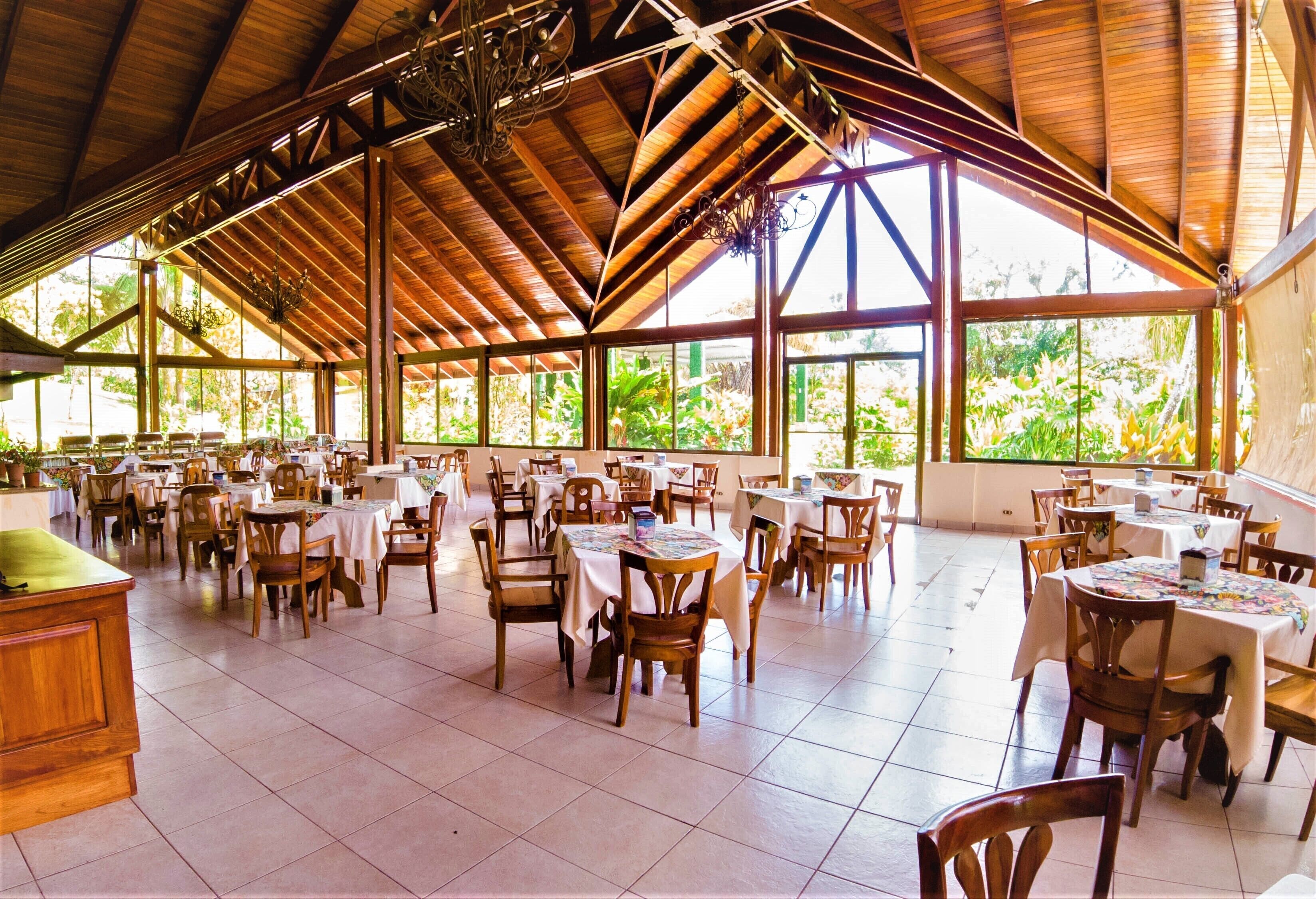 A spacious indoor dining area with high ceilings and large windows, allowing natural light to fill the space. Tables are arranged in a casual layout, and the area is furnished with wooden chairs and tables.
