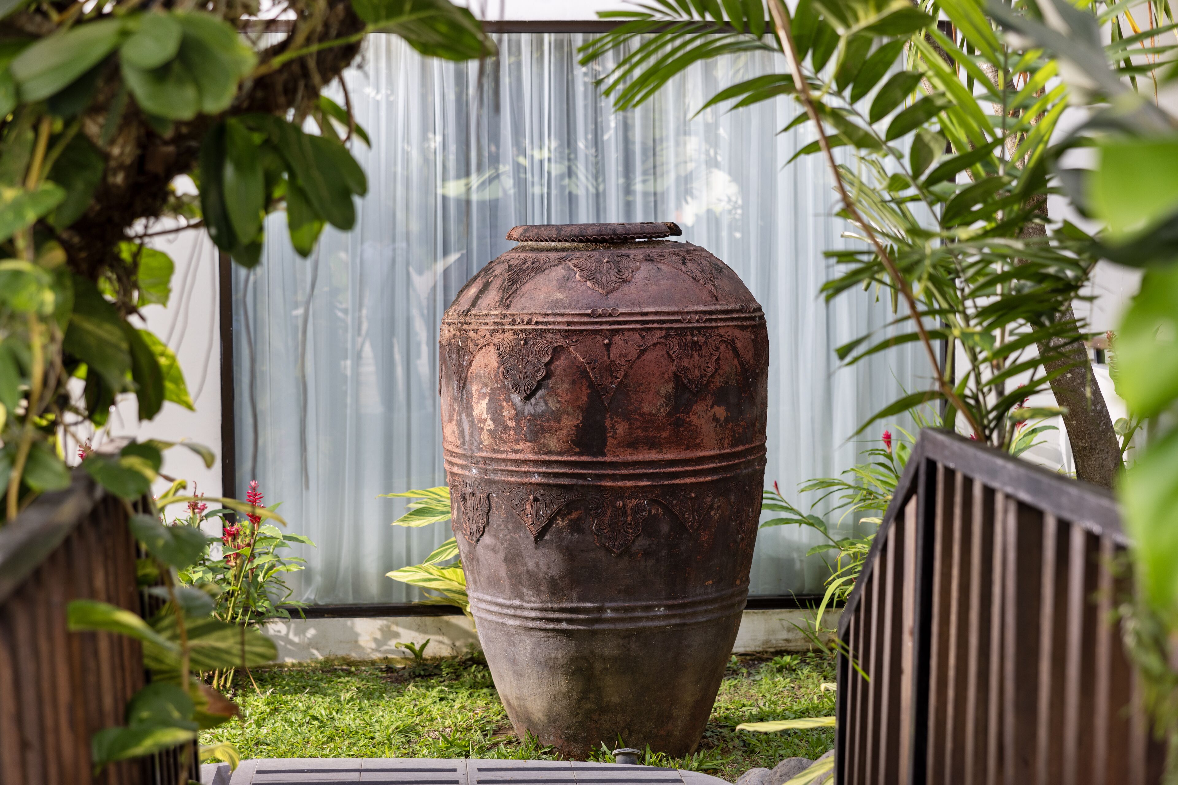 A large, rustic ceramic pot in a garden setting with a metal fence and greenery.