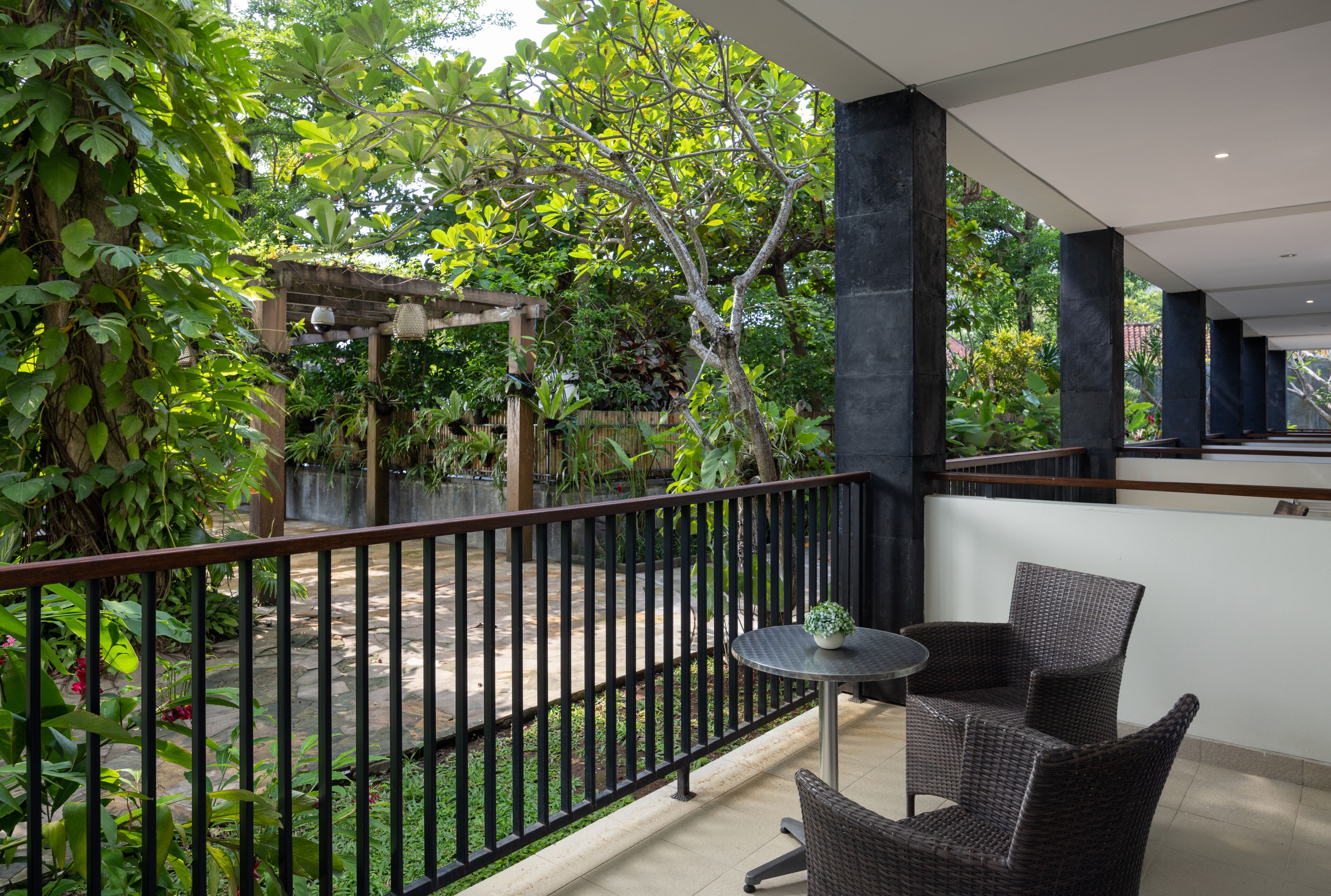 A balcony with wicker furniture, a small table, and a view of a garden with a pergola and trees.