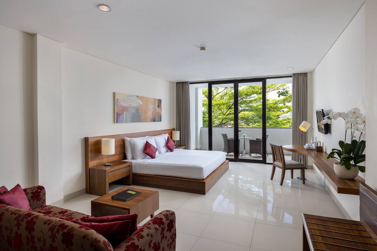 Une chambre d’hôtel moderne dotée d’un grand lit, d’un canapé, d’un bureau et d’un balcon offrant une vue sur la verdure.