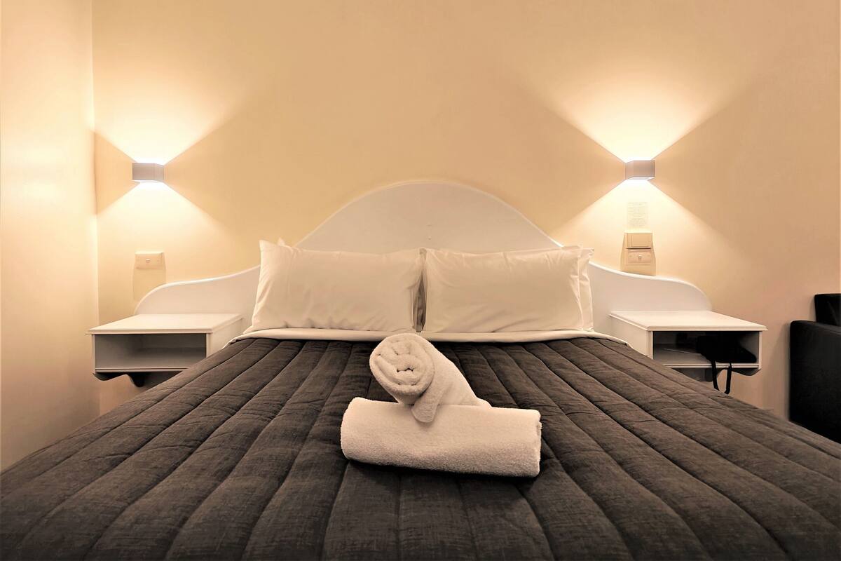 A neatly made bed with a folded towel in the center, flanked by two bedside tables with lamps, in a room with a curved headboard and light-colored walls.