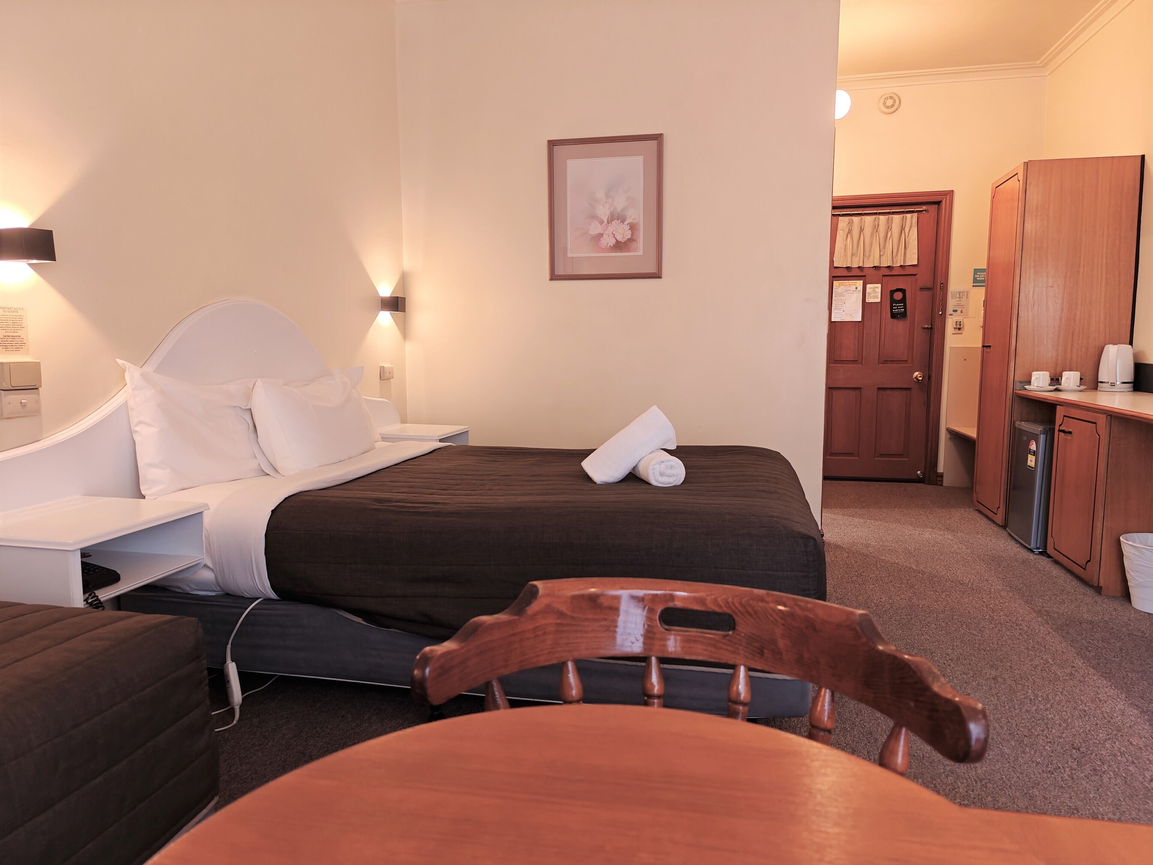 Una habitación de hotel con cama, mesita de noche, silla y una pequeña zona de kitchenette.