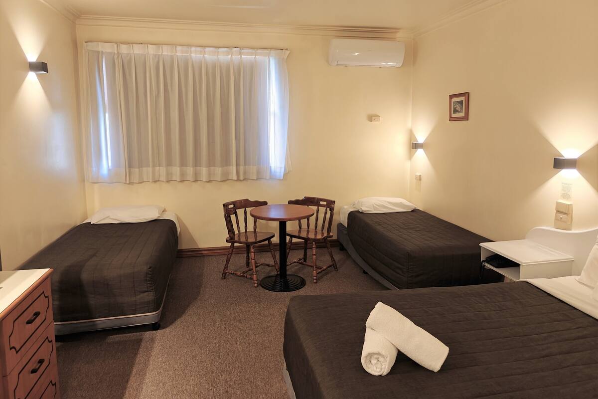 A hotel room with two single beds, a small table, and chairs. There is a window with curtains, an air conditioning unit, and a wall-mounted light fixture.