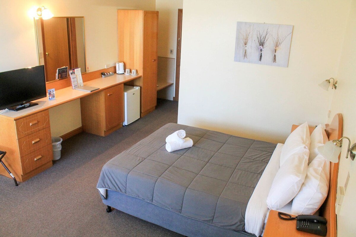A hotel room with a bed, a desk with a computer, a small fridge, a mirror, and a wall-mounted picture.