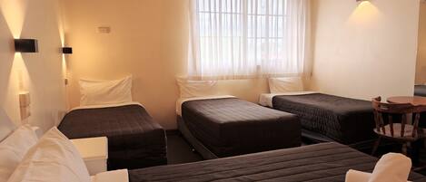 A hotel room with two single beds, a double bed, a small table, and a chair. There is an air conditioning unit on the wall and a window with sheer curtains.