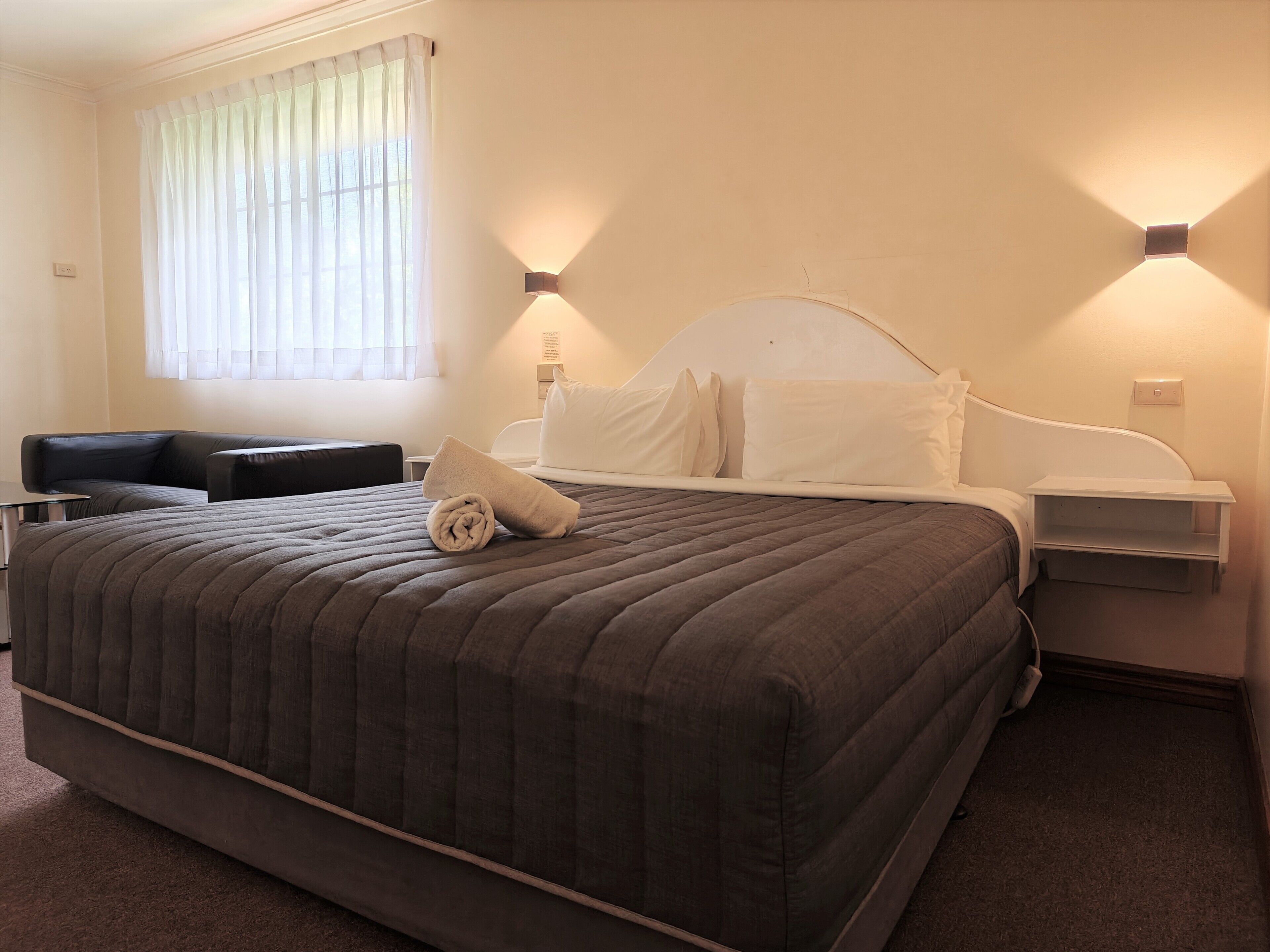 A neatly made bed with a rolled towel, a bedside table, a nightstand, and a window with sheer curtains.