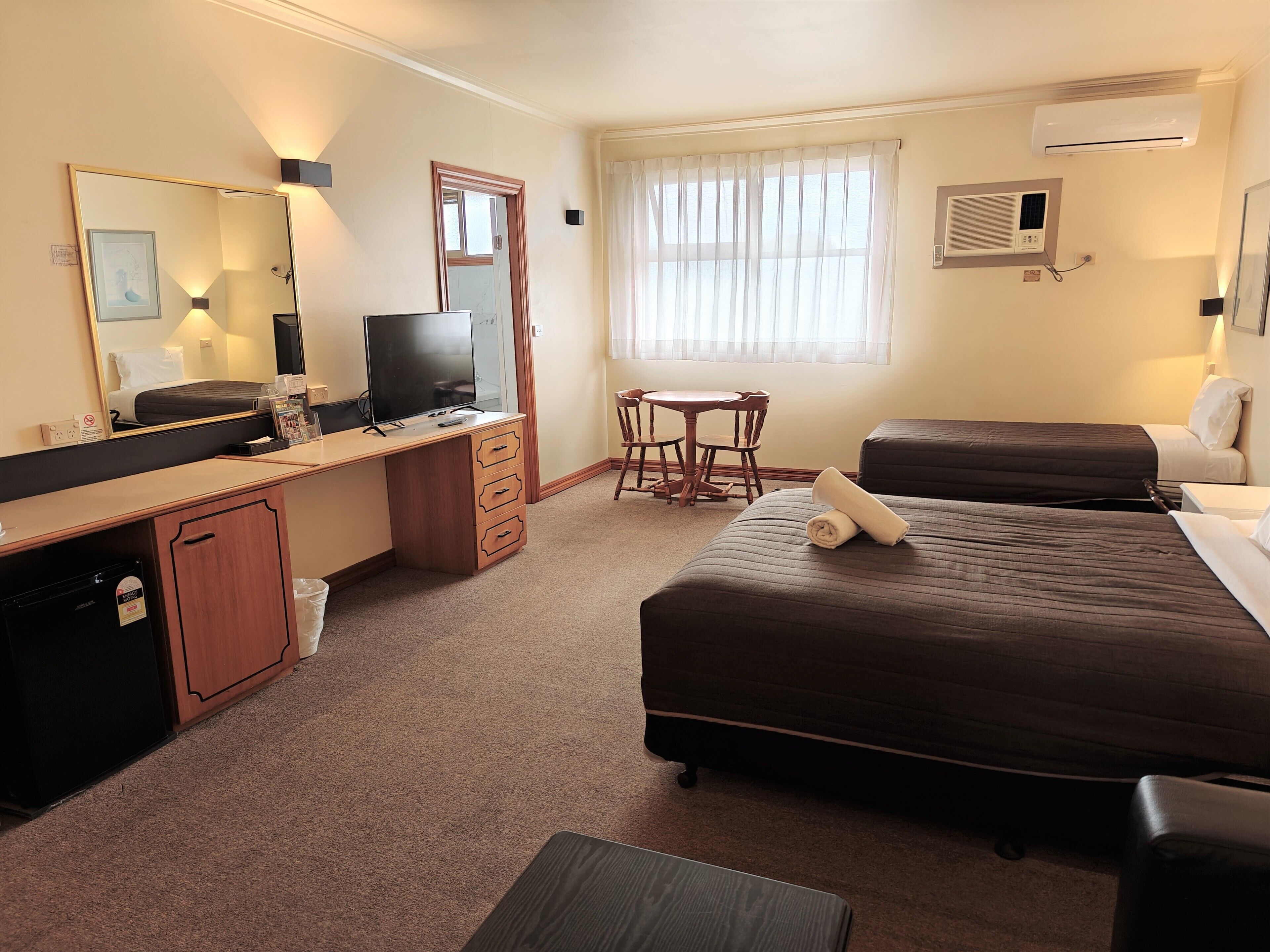 A hotel room with a bed, a desk with a computer, a chair, a small table, and a window with curtains.