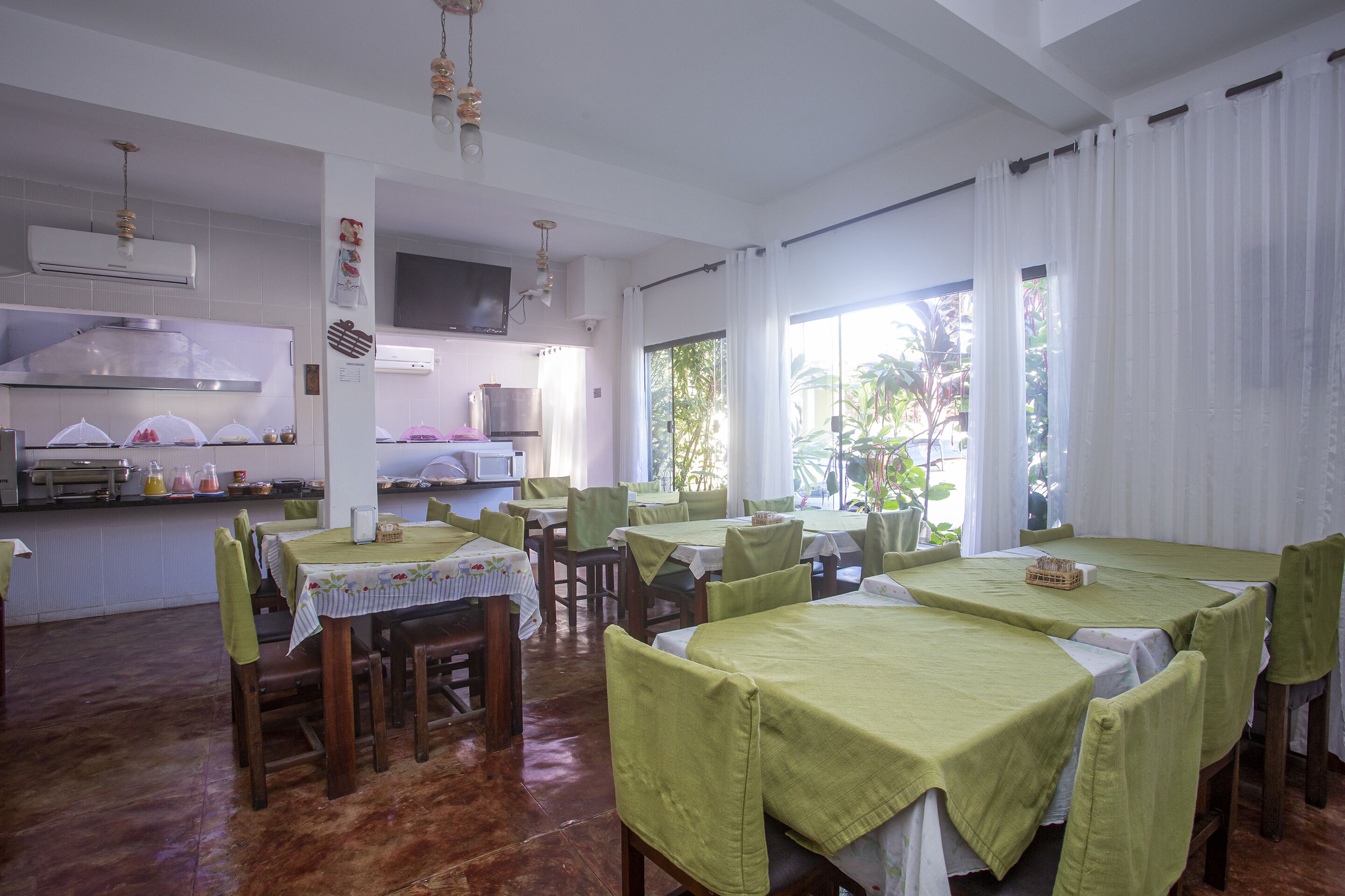 Un comedor con mesas cubiertas con manteles verdes, sillas de madera y una cocina al fondo.