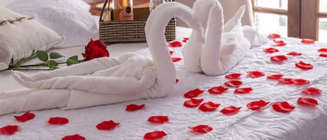 A bed with a white quilt, red rose petals, and two white towels shaped like swans.