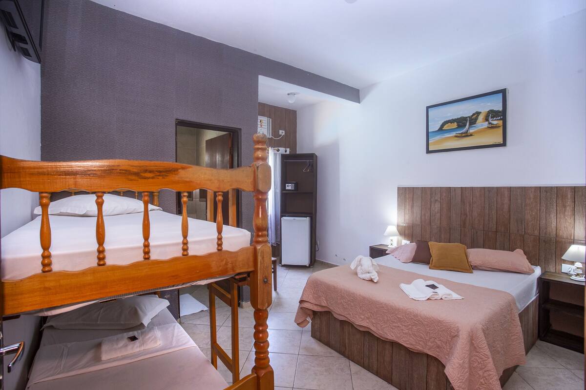 Ein Hotelzimmer mit Etagenbett, einem Einzelbett, einem Holzkopfende, einem kleinen Kühlschrank, einem Gemälde einer Strandansicht und einer Wandpaneele aus Holz.