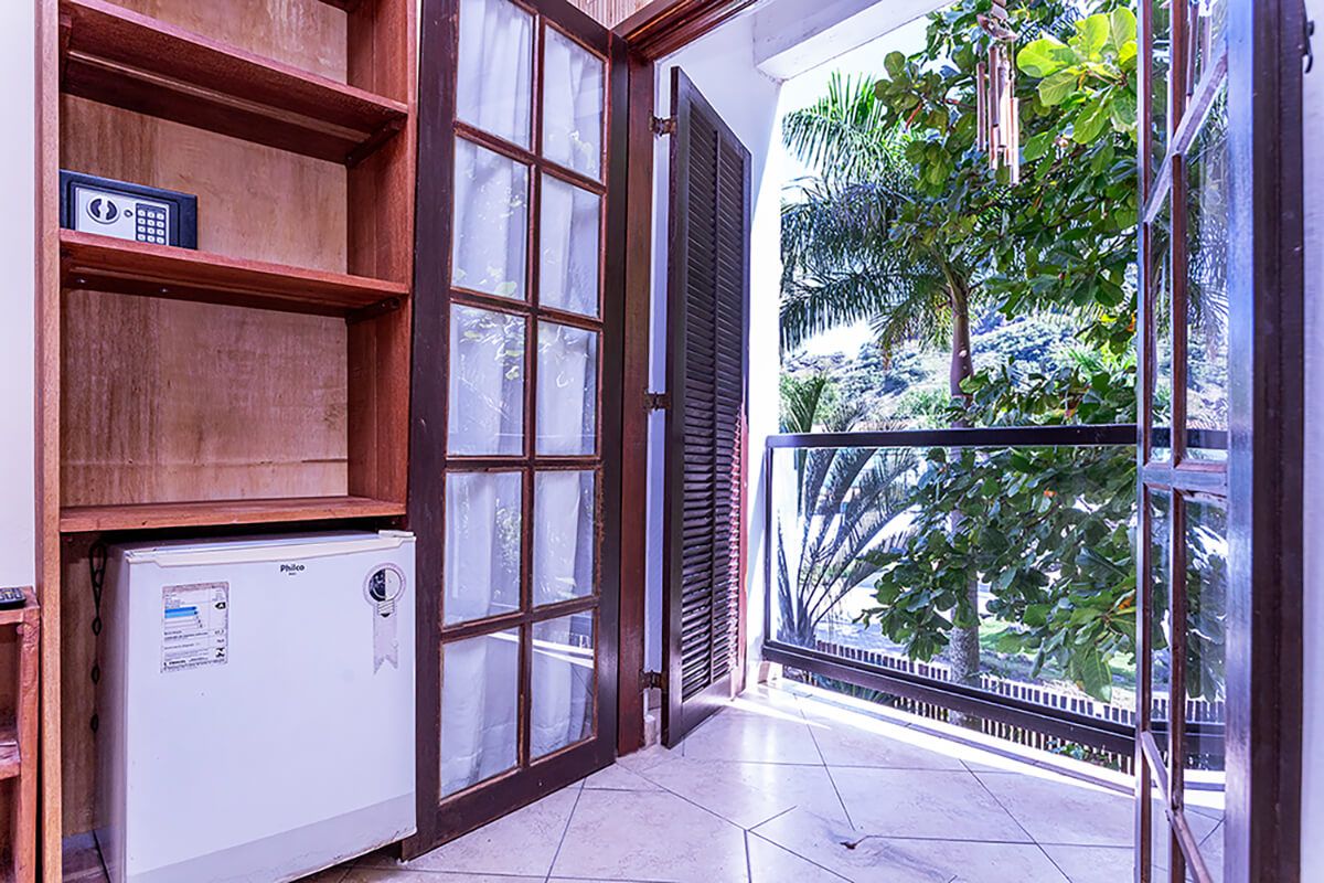 A room with a refrigerator, wooden shelves, a window with a view of greenery, and a balcony with a railing.