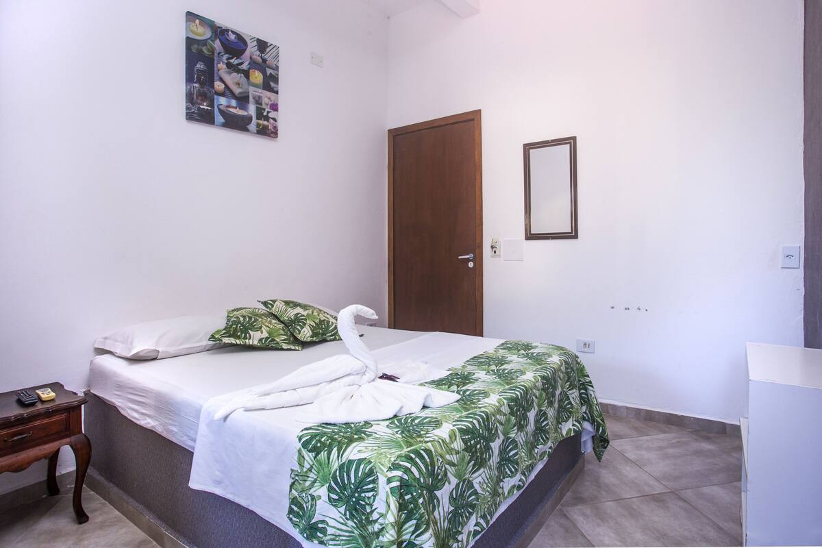 Ein ordentlich eingerichtetes Hotelzimmer mit einem Bett, einem kleinen Nachttisch, einem wandmontierten Spiegel und einer Tür.