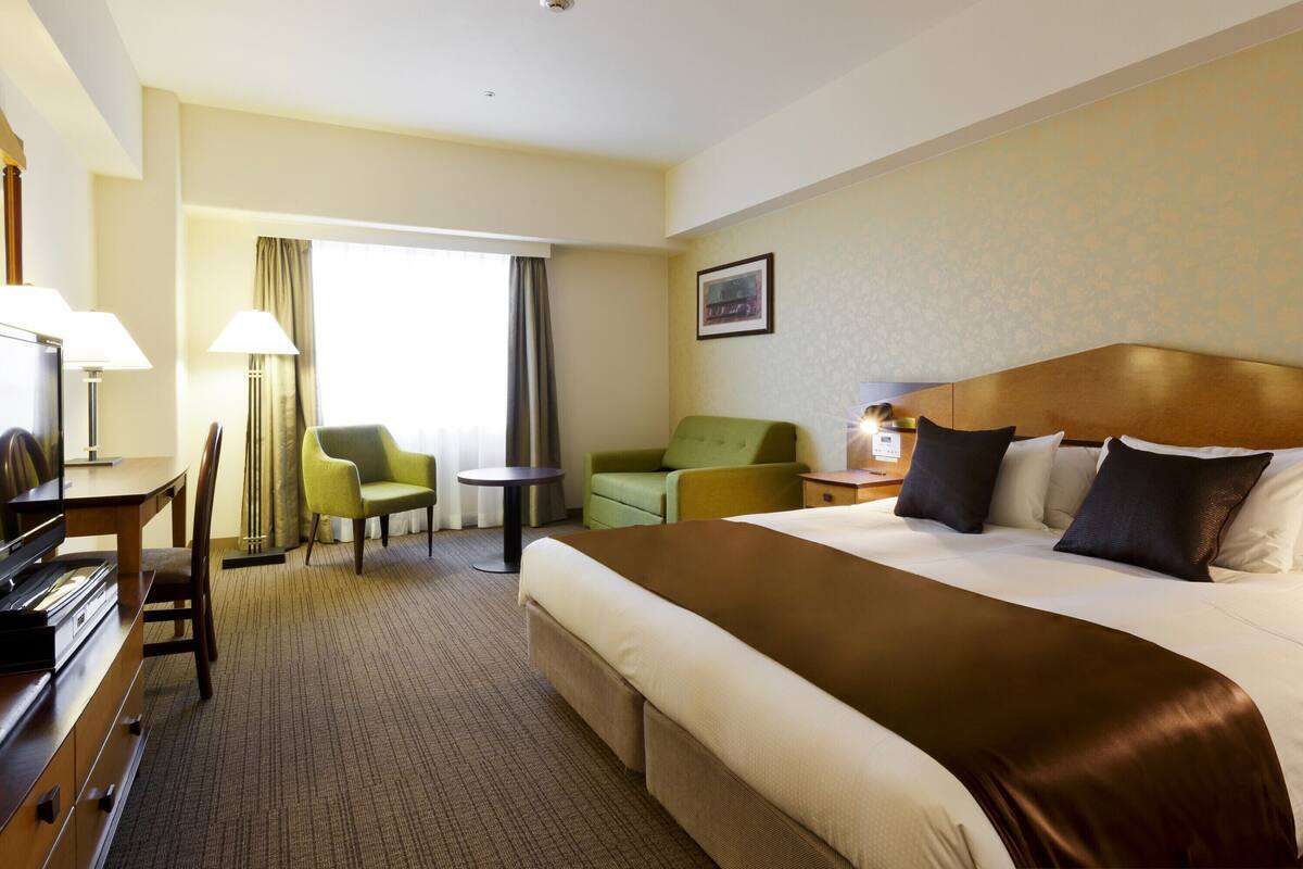 A hotel room with a large bed, a desk with a lamp, a chair, a small table, and a window with curtains.