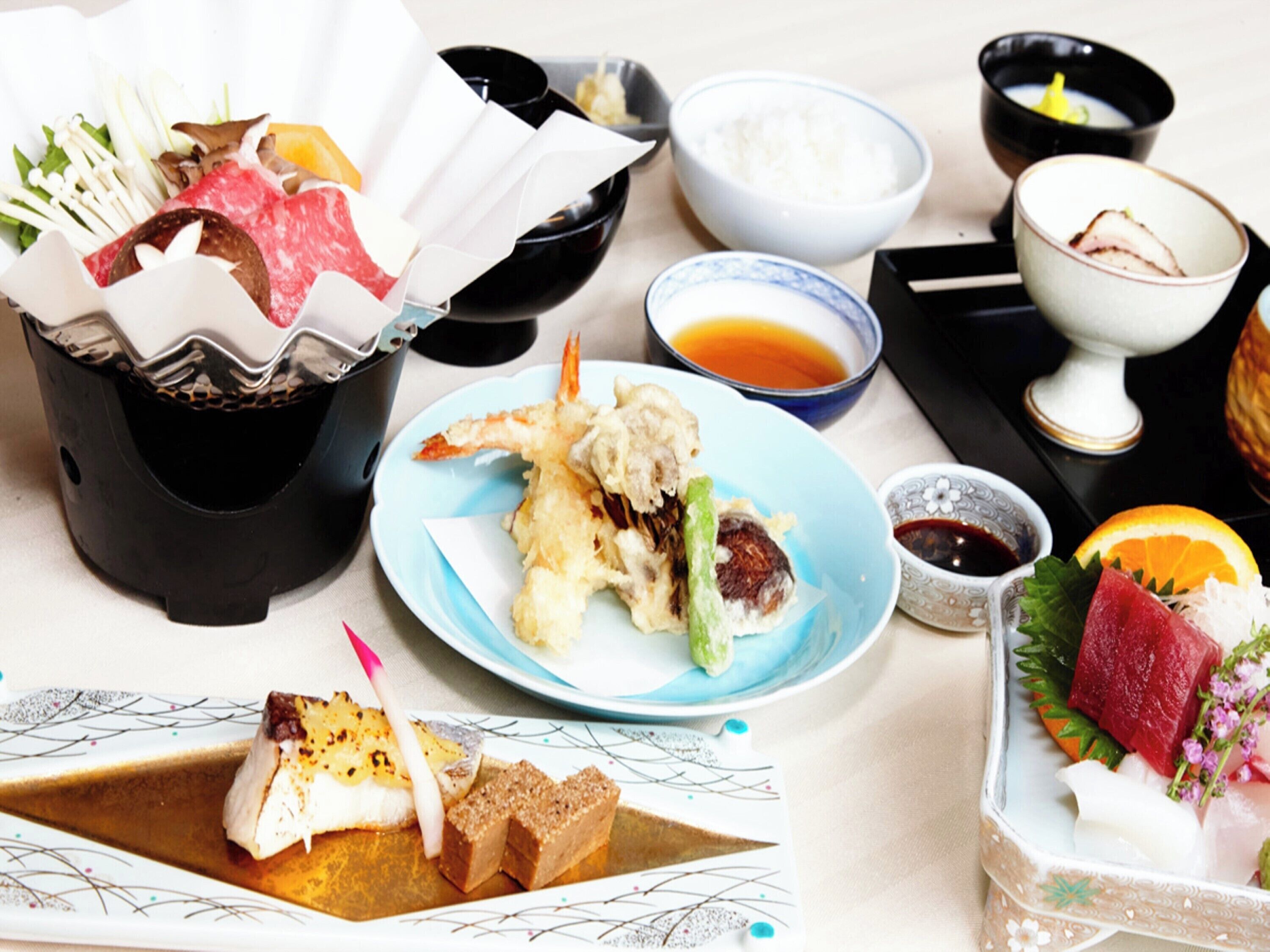 A variety of Japanese dishes including sashimi, tempura, and a selection of vegetables and meats, served on a table with multiple bowls and plates.