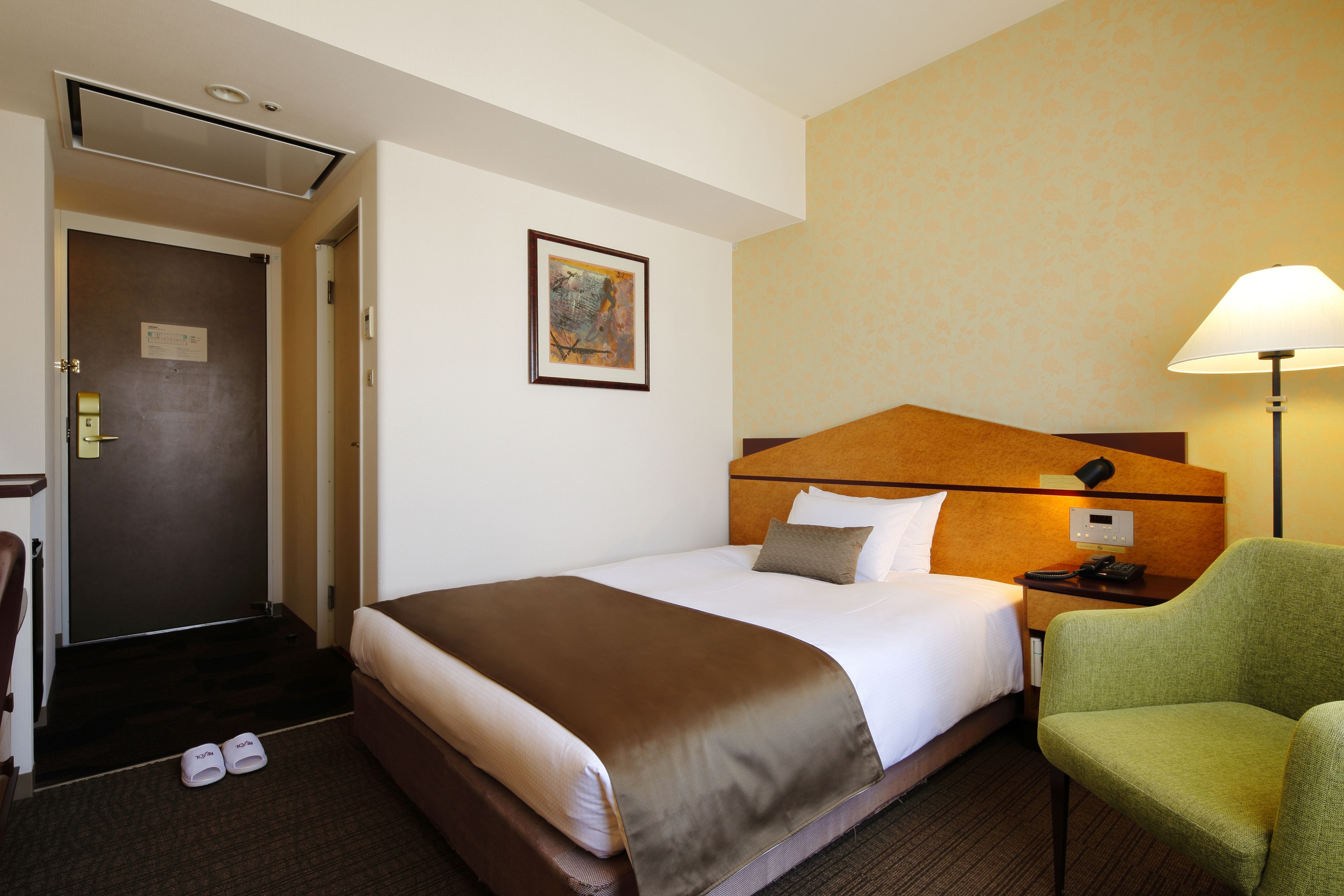A hotel room with a bed, a green chair, a nightstand with a lamp, and a door.