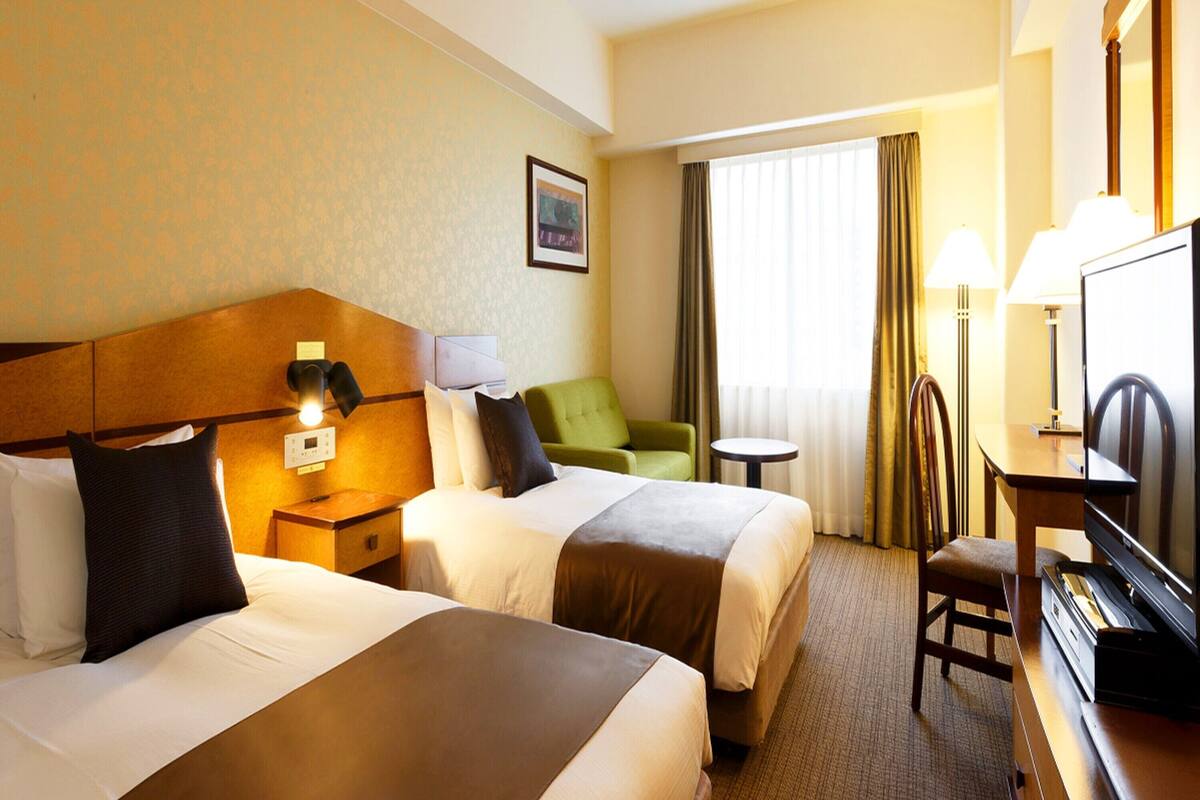 A hotel room with two beds, a desk with a television, a chair, and a lamp.