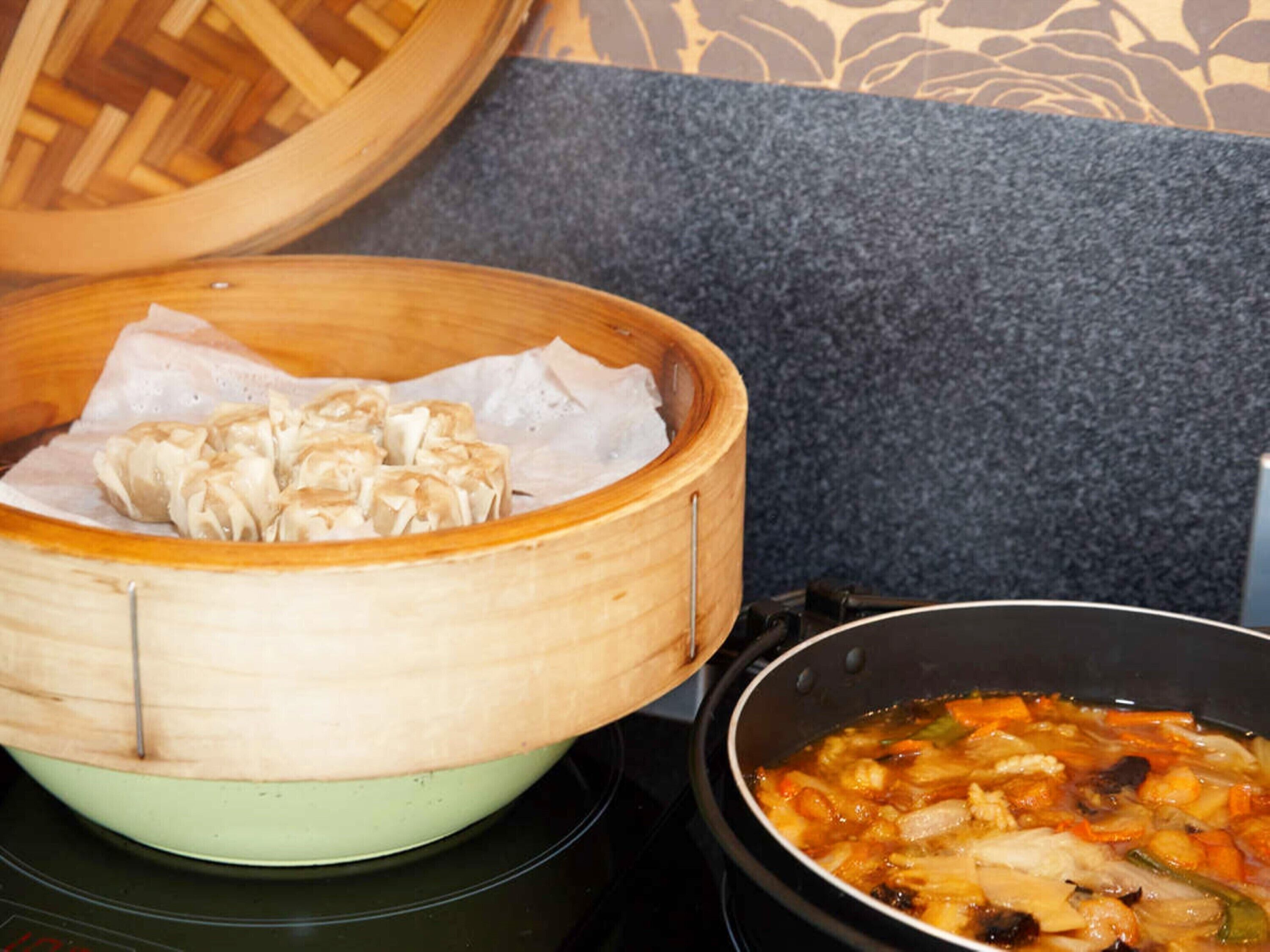A bamboo steamer with dumplings on a stove, next to a pot of soup.