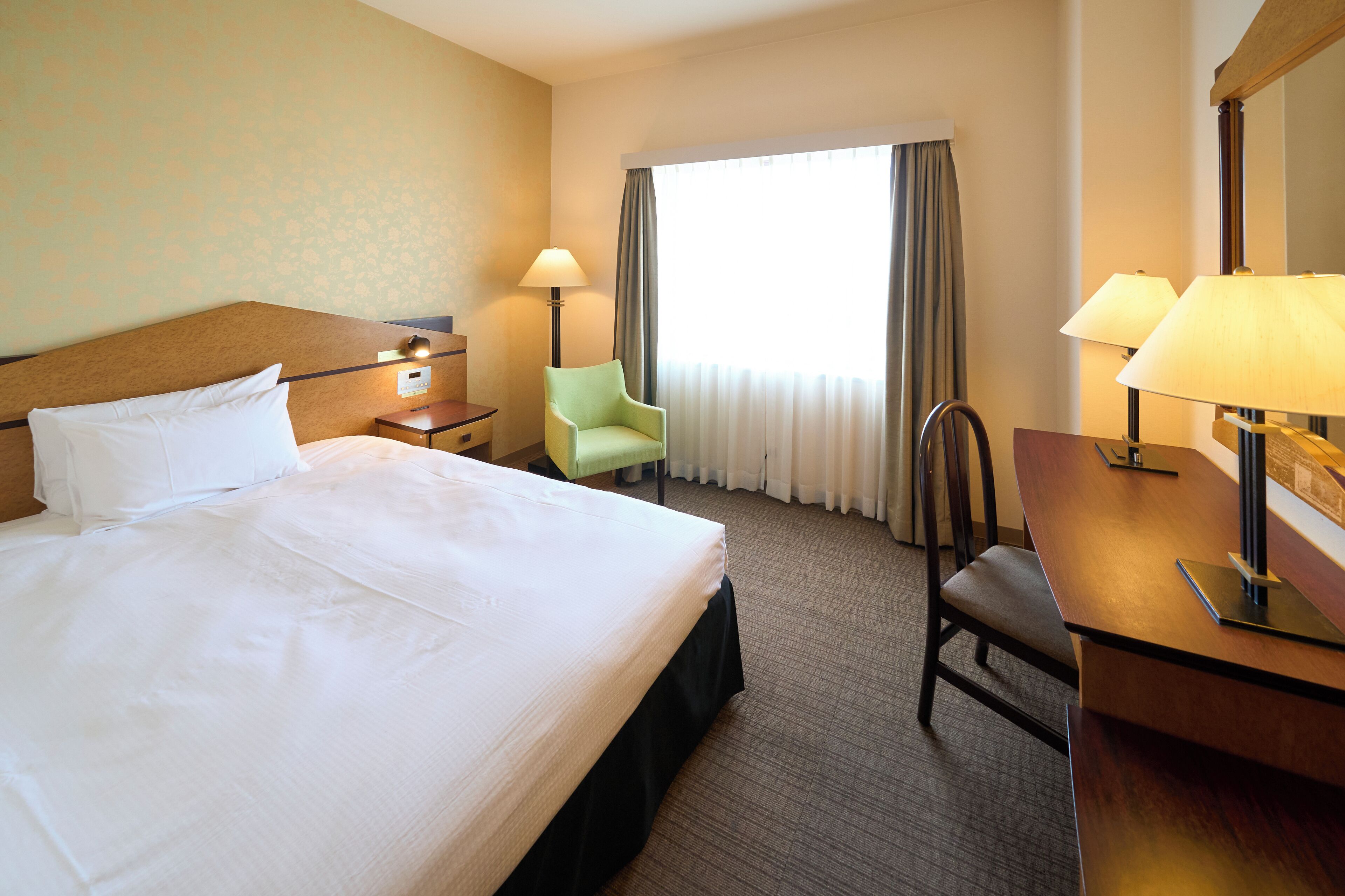 Quarto de hotel com uma cama grande, uma escrivaninha, uma cadeira, um abajur e uma janela com cortinas.