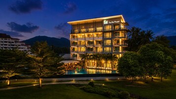 A multi-story hotel with a pool in the foreground, surrounded by greenery and a mountainous backdrop.
