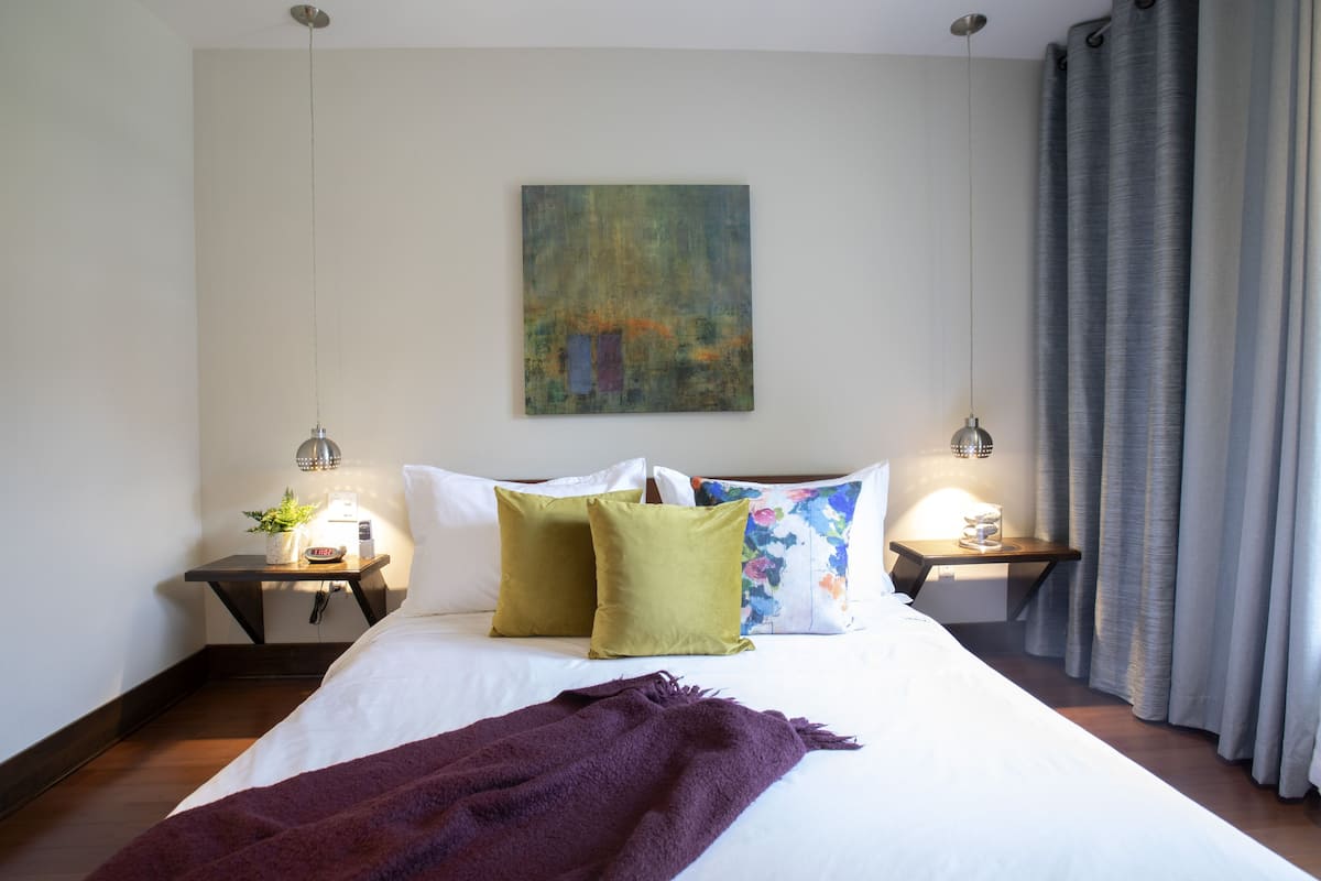Una cama bien hecha con una manta morada, dos mesitas de noche con lámparas, un cojín floral y un cuadro en la pared.