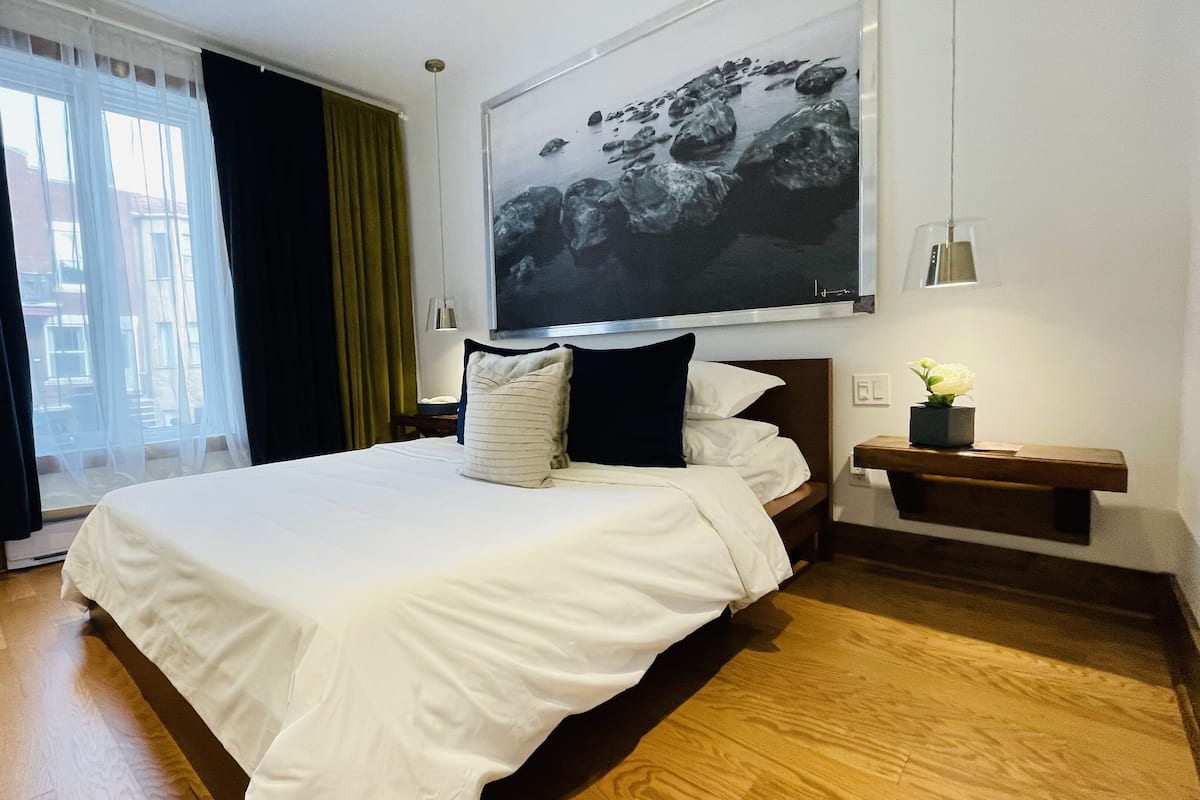 Habitación de hotel con una cama grande, una mesita de noche con un jarrón de flores, una ventana con cortinas y una obra de arte colgada en la pared.