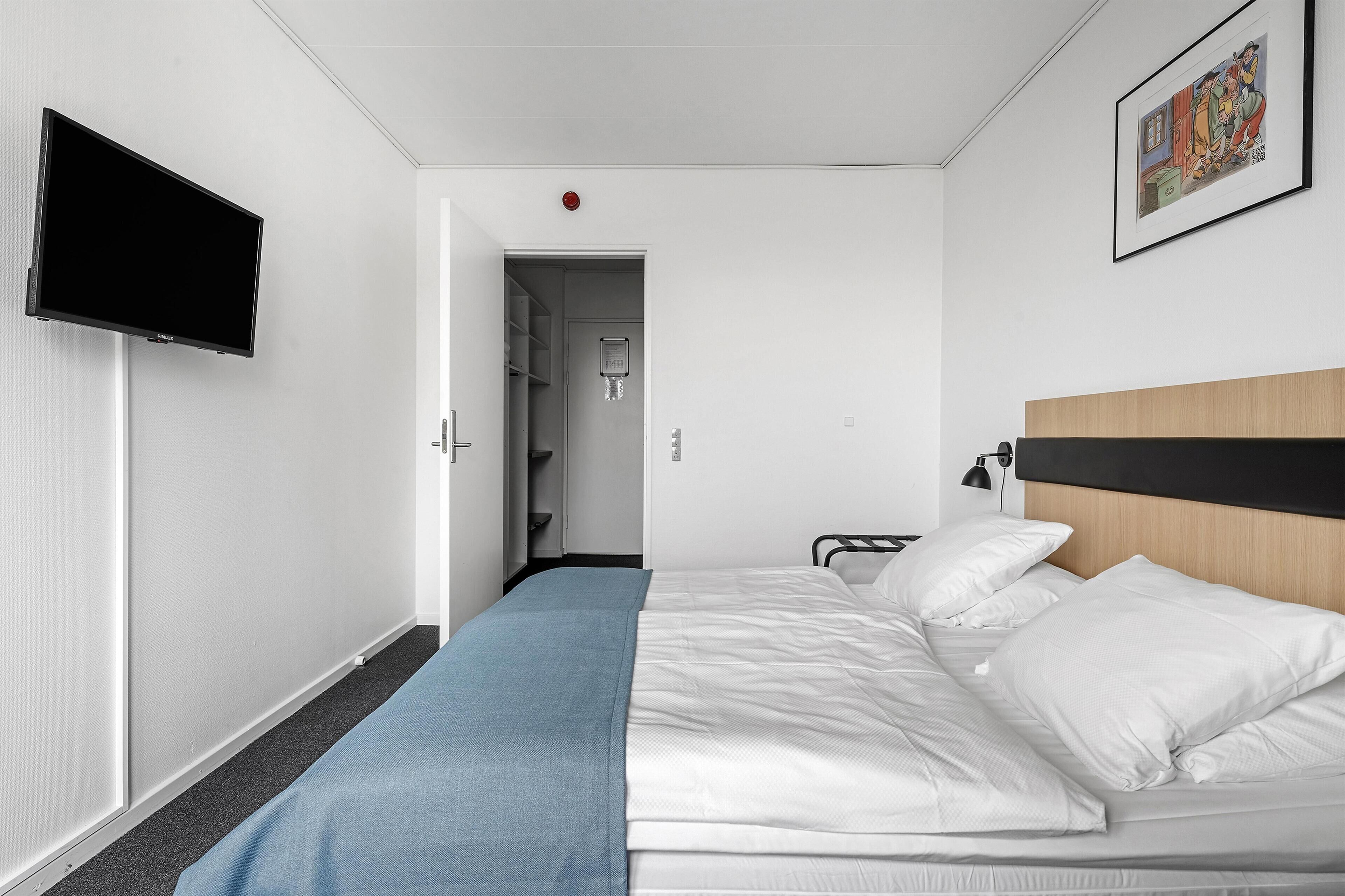 A hotel room with a bed, a TV mounted on the wall, a door leading to a bathroom, and a framed picture on the wall.