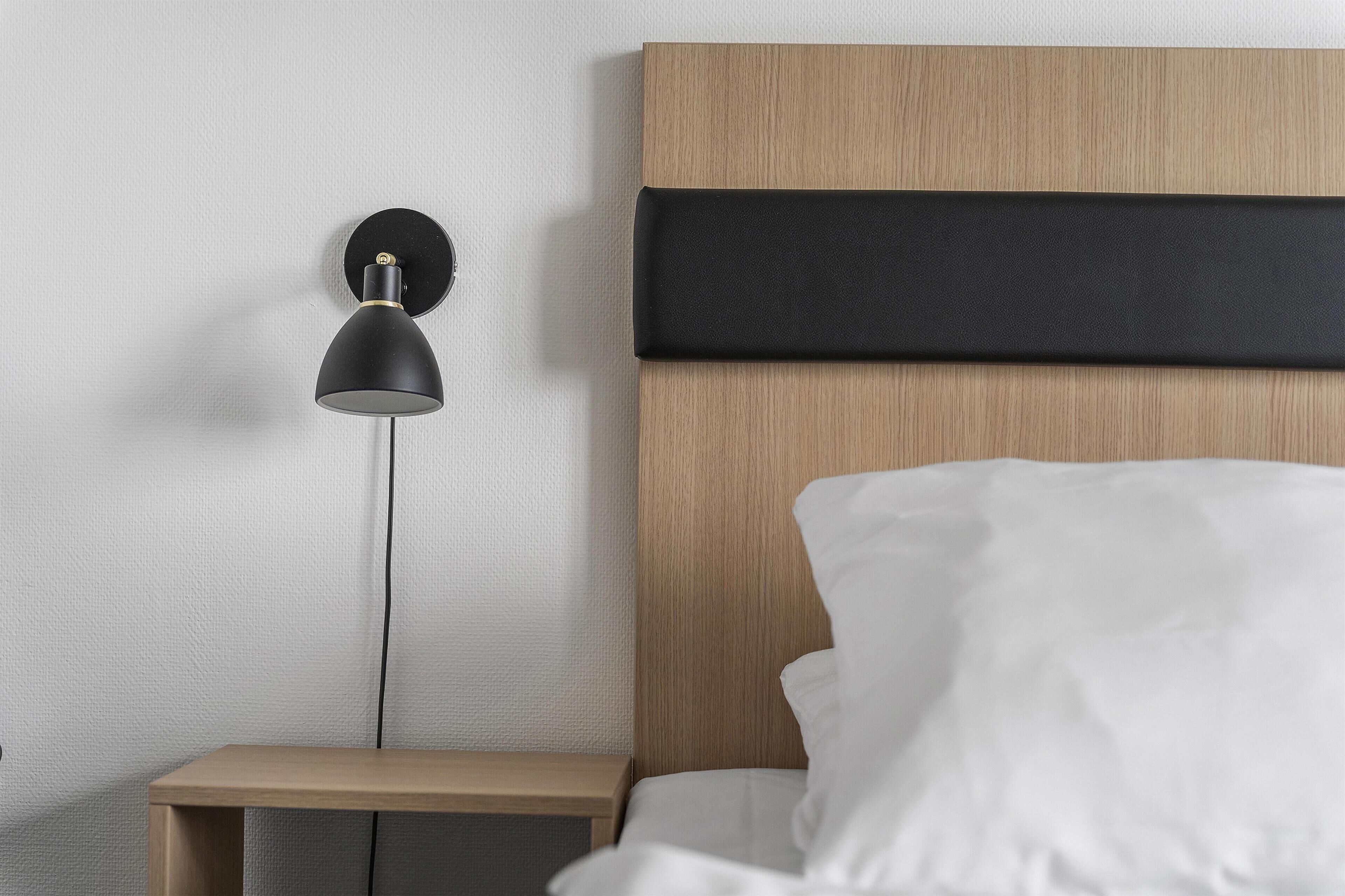 Una cama con ropa de cama blanca, cabecera de madera, una lámpara negra sobre una mesita de noche y una pared lisa.